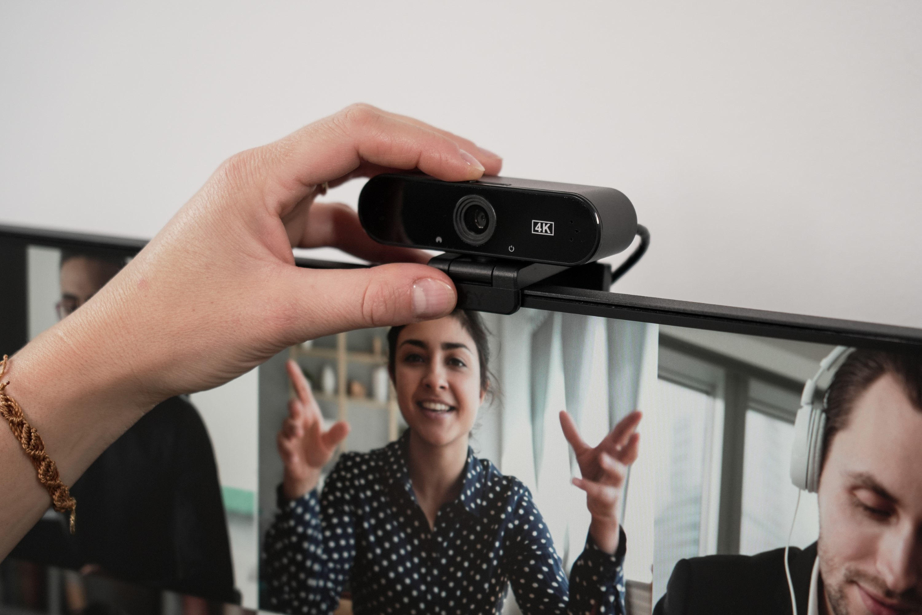 Dłoń kobiety regulująca czarną kamerę internetową zamontowaną na monitorze wyświetlającym wideokonferencję.