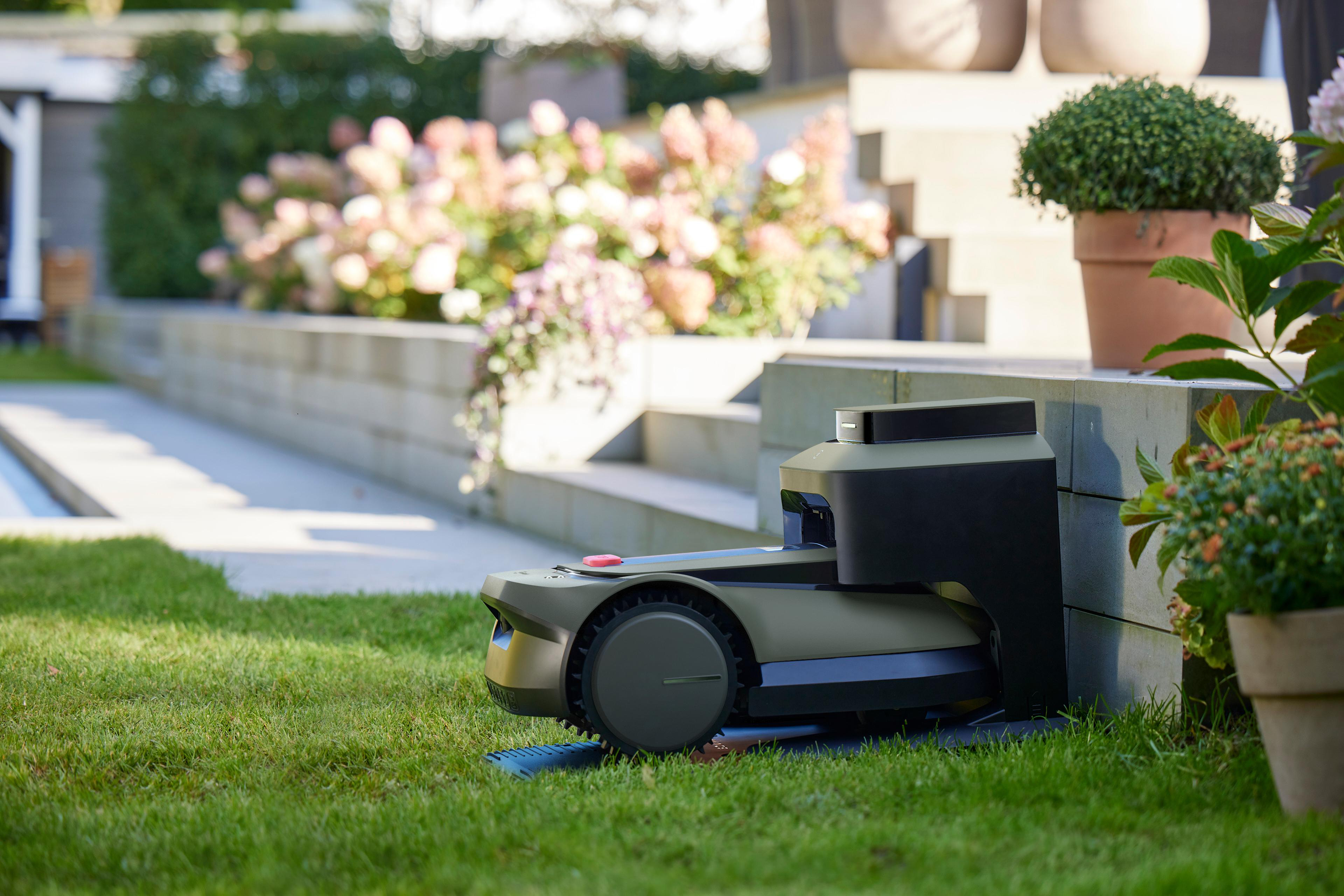 Grüner Roboter-Rasenmäher auf Gras, in der Nähe einer Betonstruktur und Pflanzen.