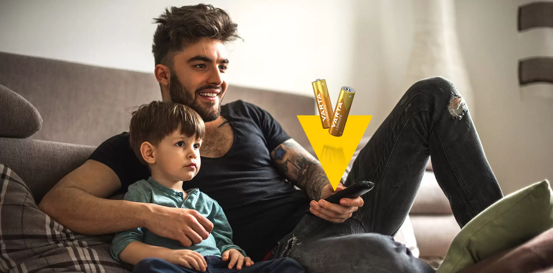 Apa és fia tévéznek, elemekkel és távirányítóval az előtérben.