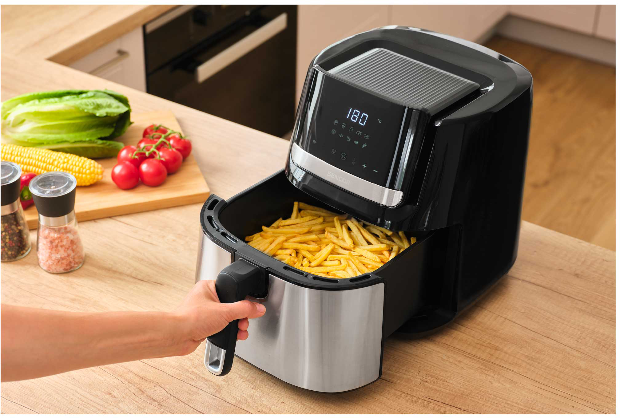 Egy air fryer sült krumplival, egy kéz a fogantyút tartja, fa felületen.