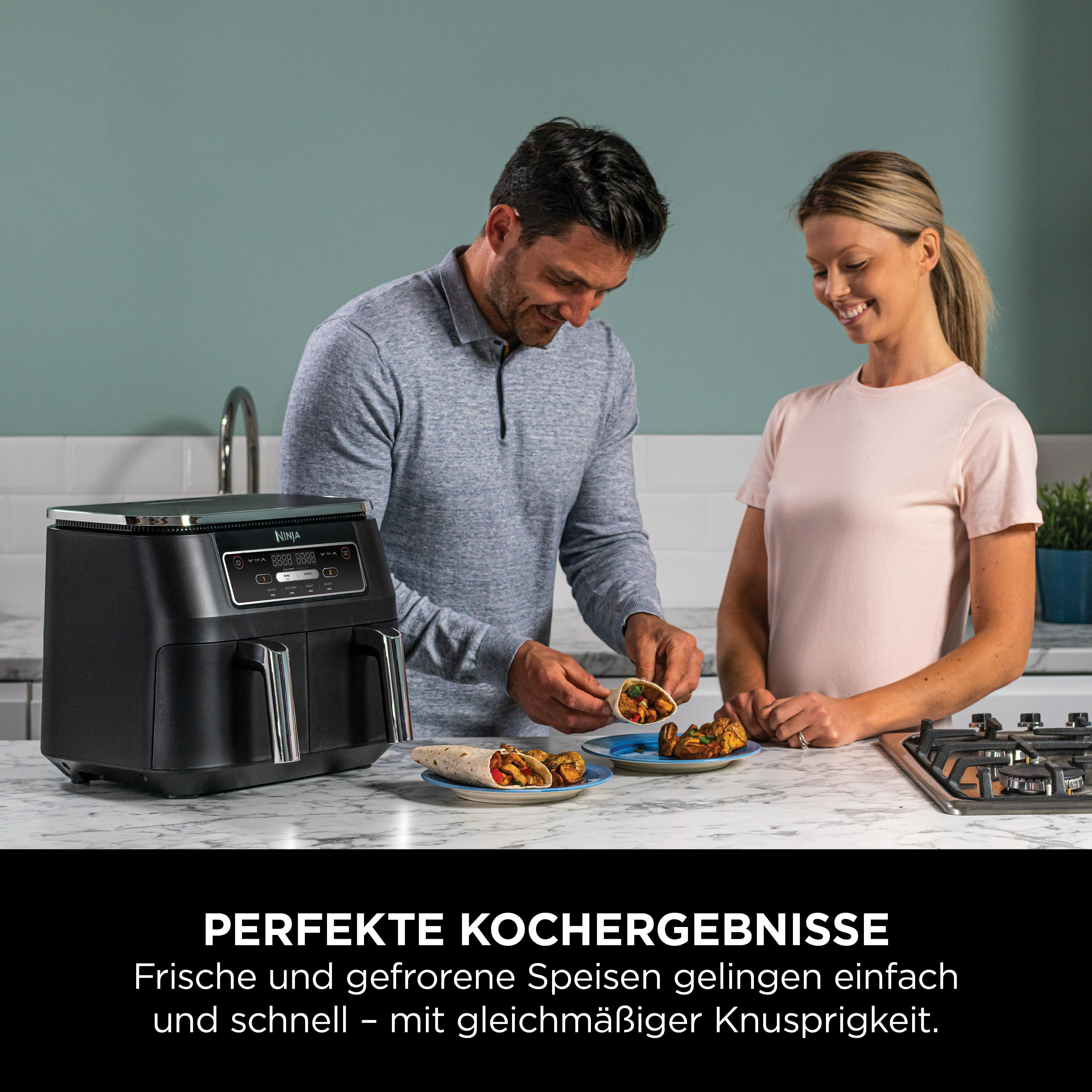 Ein Mann und eine Frau kochen mit einer schwarzen Heißluftfritteuse. Essen ist auf Tellern. Sie lächeln.