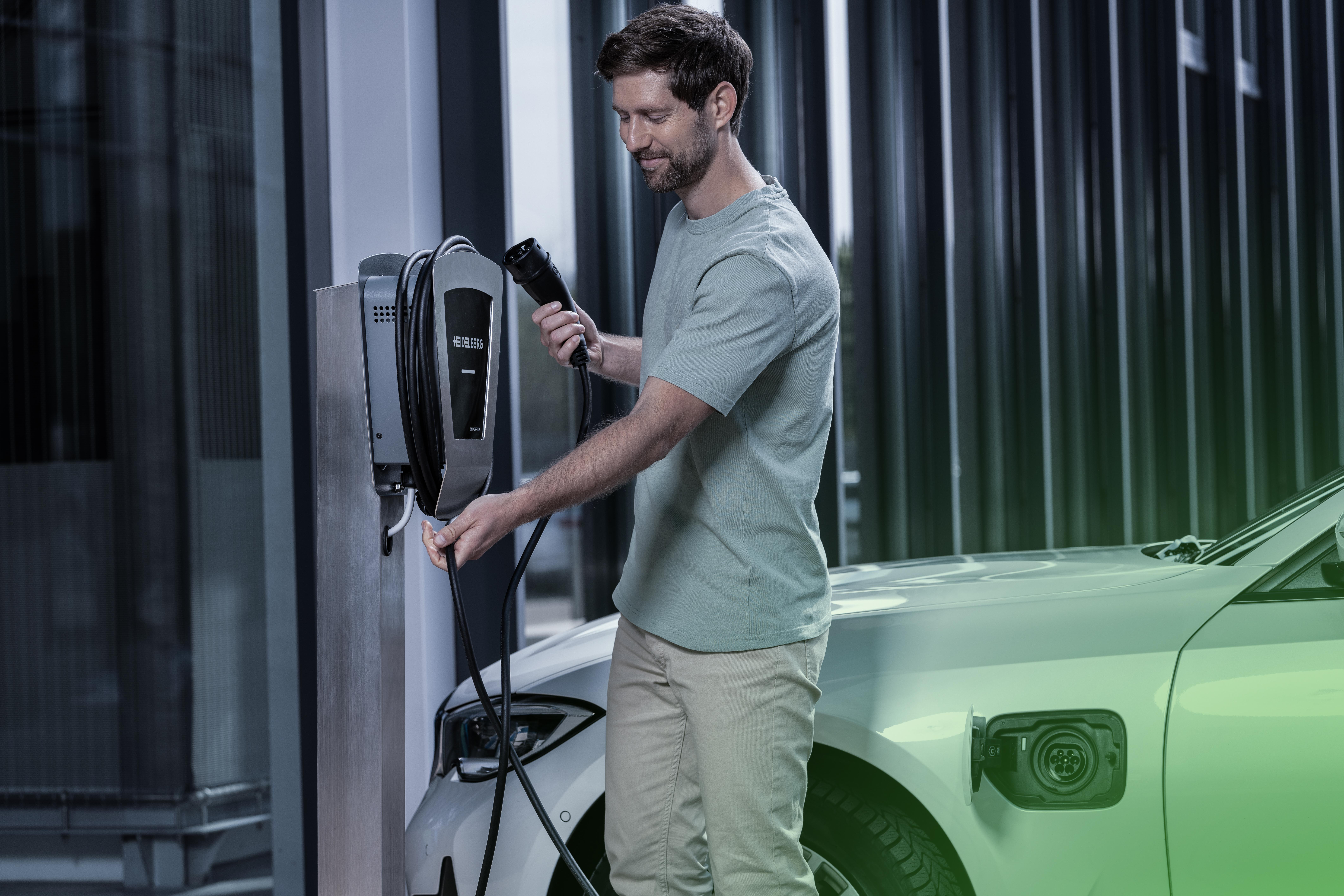 Ein Mann lädt ein Auto auf. Eine Ladestation befindet sich links, ein weißes Auto rechts.