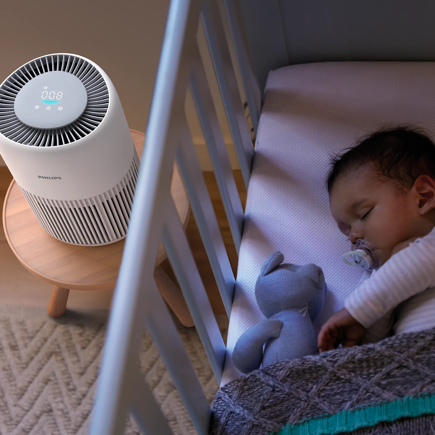 Un bébé dort dans un berceau à côté d'un purificateur d'air blanc. Un ours en peluche est dans le berceau.