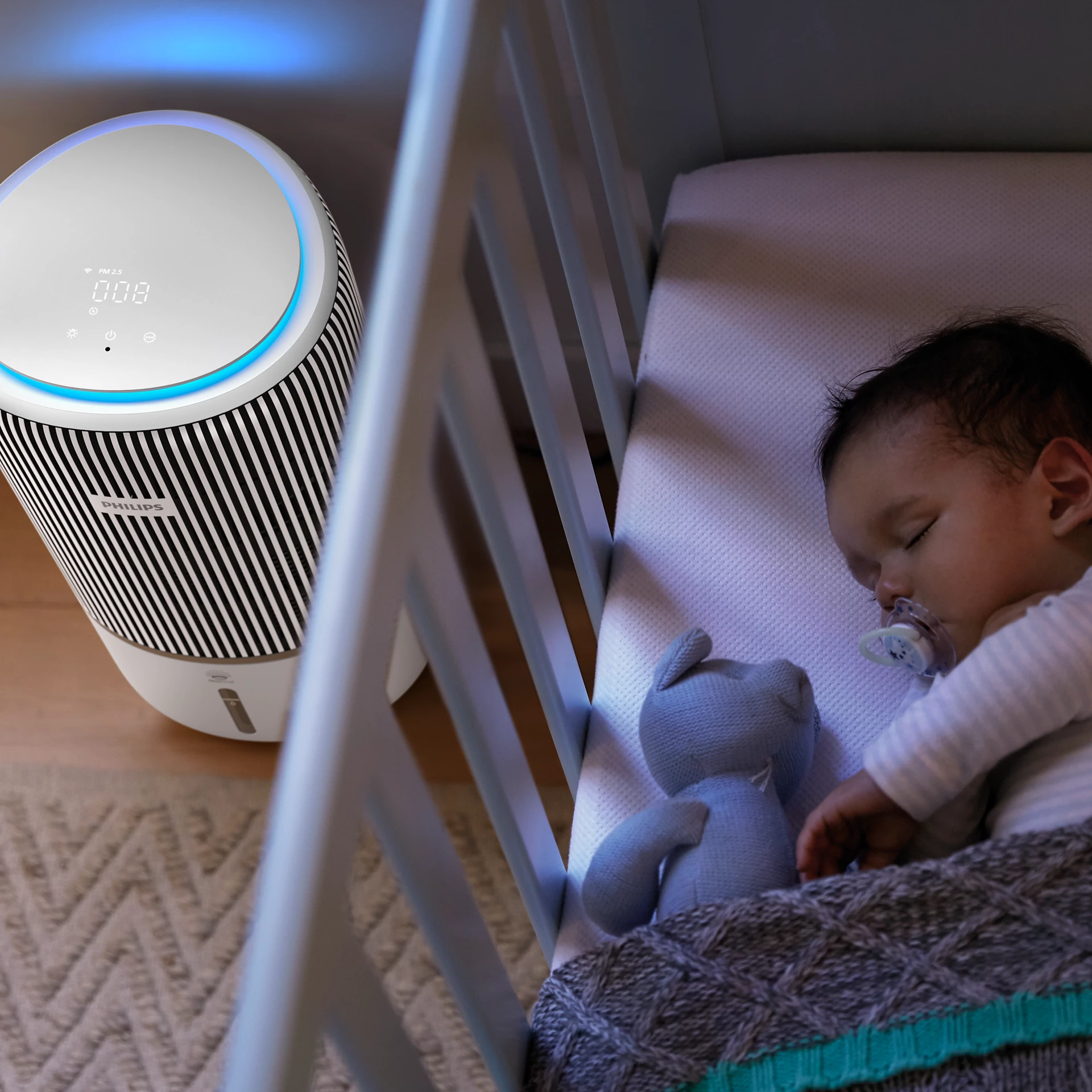 Un bébé dort dans un berceau à côté d'un purificateur d'air, avec un ours en peluche.