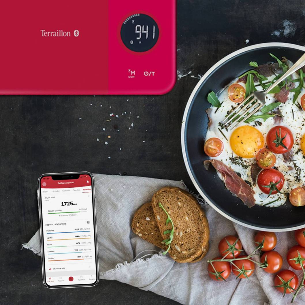 Rote Küchenwaage mit Digitalanzeige und ein Teller mit Spiegeleiern, Tomaten und Brot auf einem Tisch.