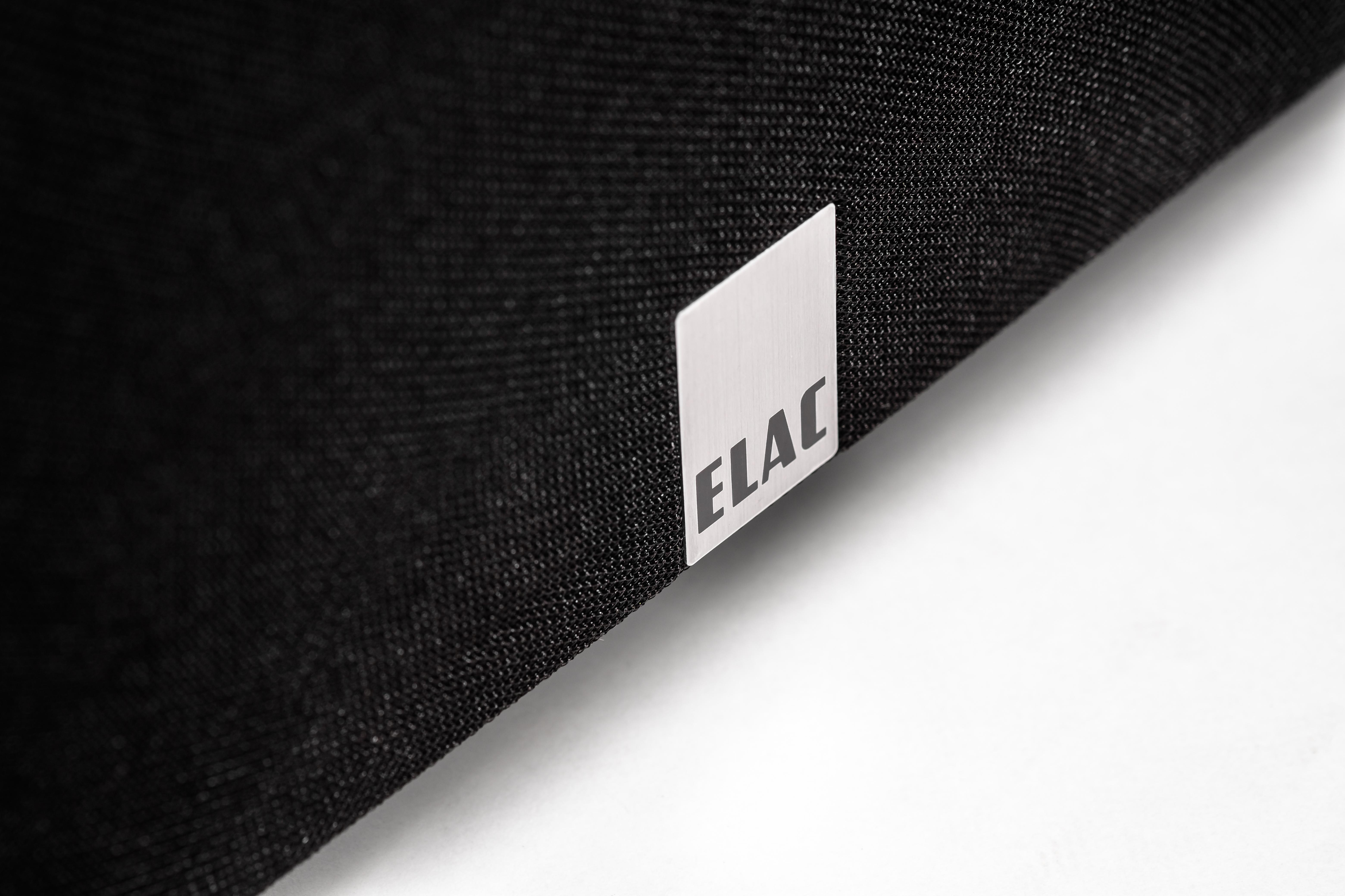 Nahaufnahme eines schwarzen Lautsprechers mit ELAC-Logo auf einer silbernen Platte. Weißer Hintergrund.