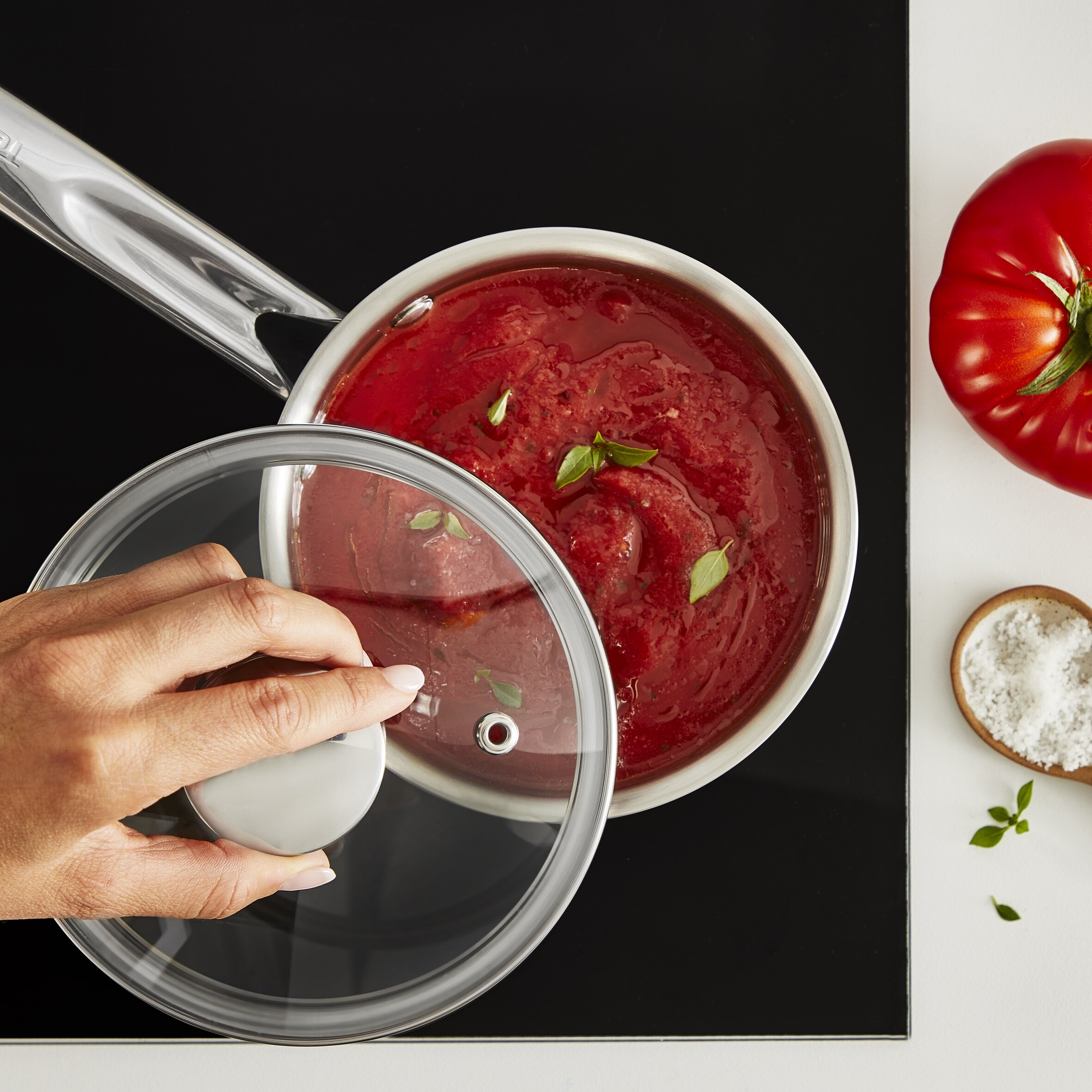 Ręka trzymająca przezroczystą pokrywkę nad garnkiem z sosem pomidorowym.