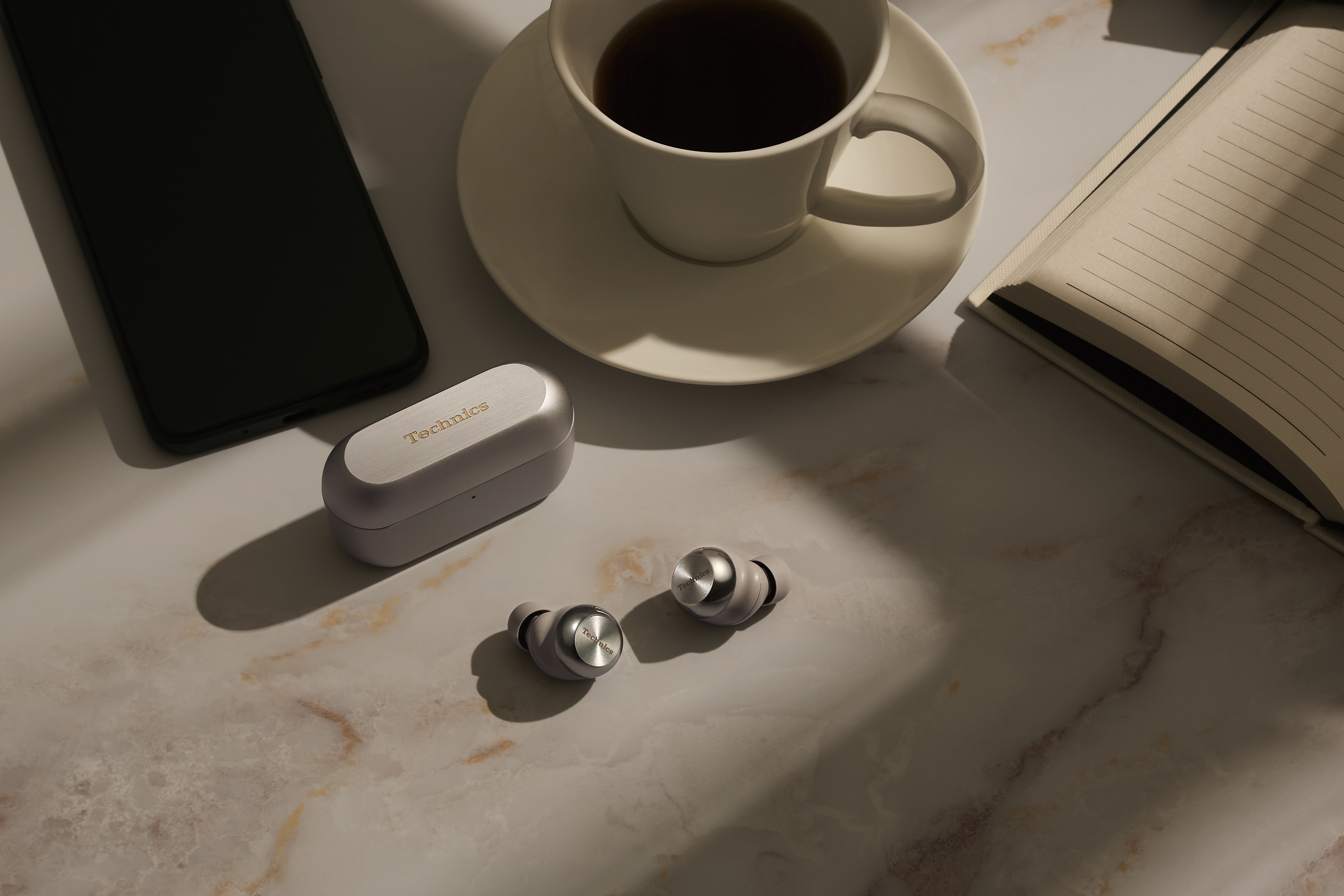 Kabellose Ohrhörer und ein Ladecase auf Marmor. Ein Smartphone und eine Tasse sind in der Nähe.