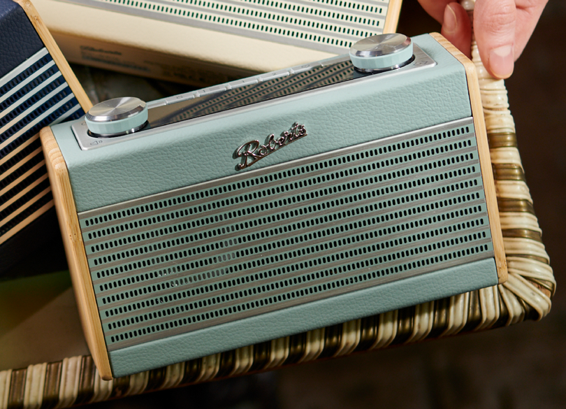 Ein türkisfarbenes Roberts-Radio auf einem Korbtisch, mit einer Hand an der Ecke. Andere Radios im Hintergrund.