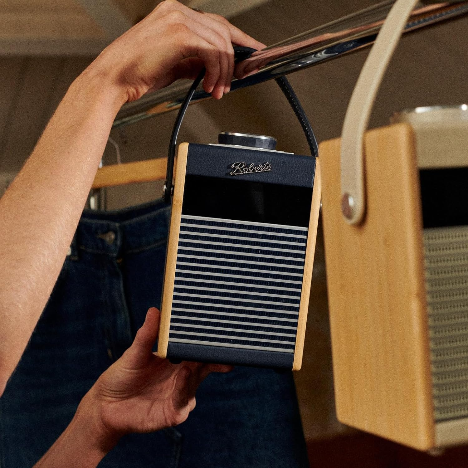 Hände halten ein marineblaues Radio mit Holzakzenten. Ein anderes Radio ist im Hintergrund. Jeans sind ebenfalls sichtbar.