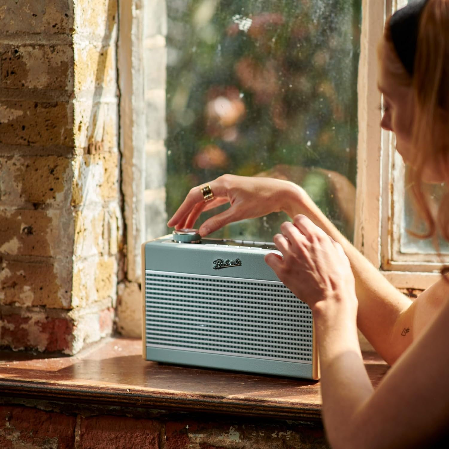 Frau, die ein Retro-Radio auf einer Fensterbank einstellt. Das Fenster zeigt eine verschwommene Szene im Freien.