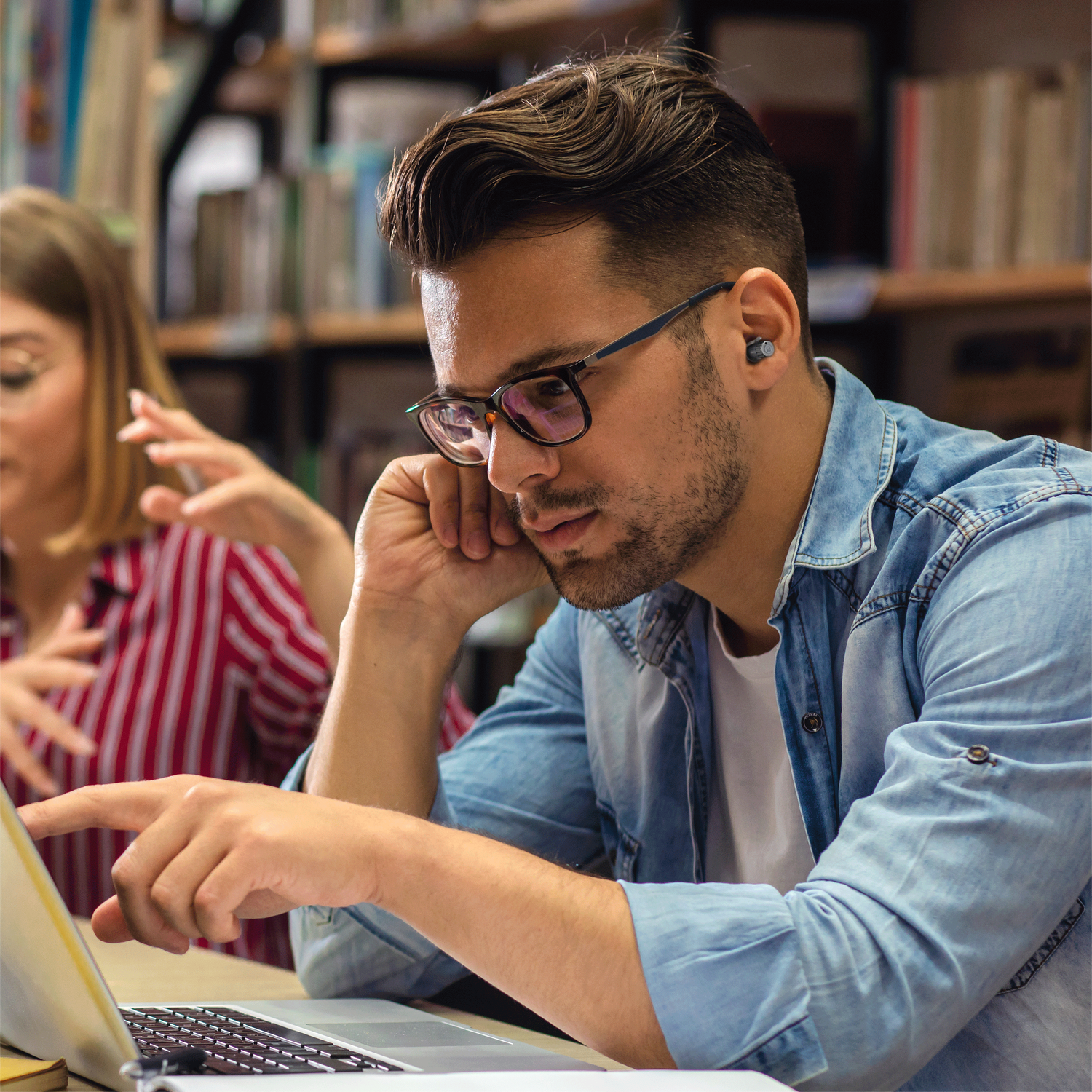 Man with glasses using a laptop. Another person in the background. Bookshelves in the background.