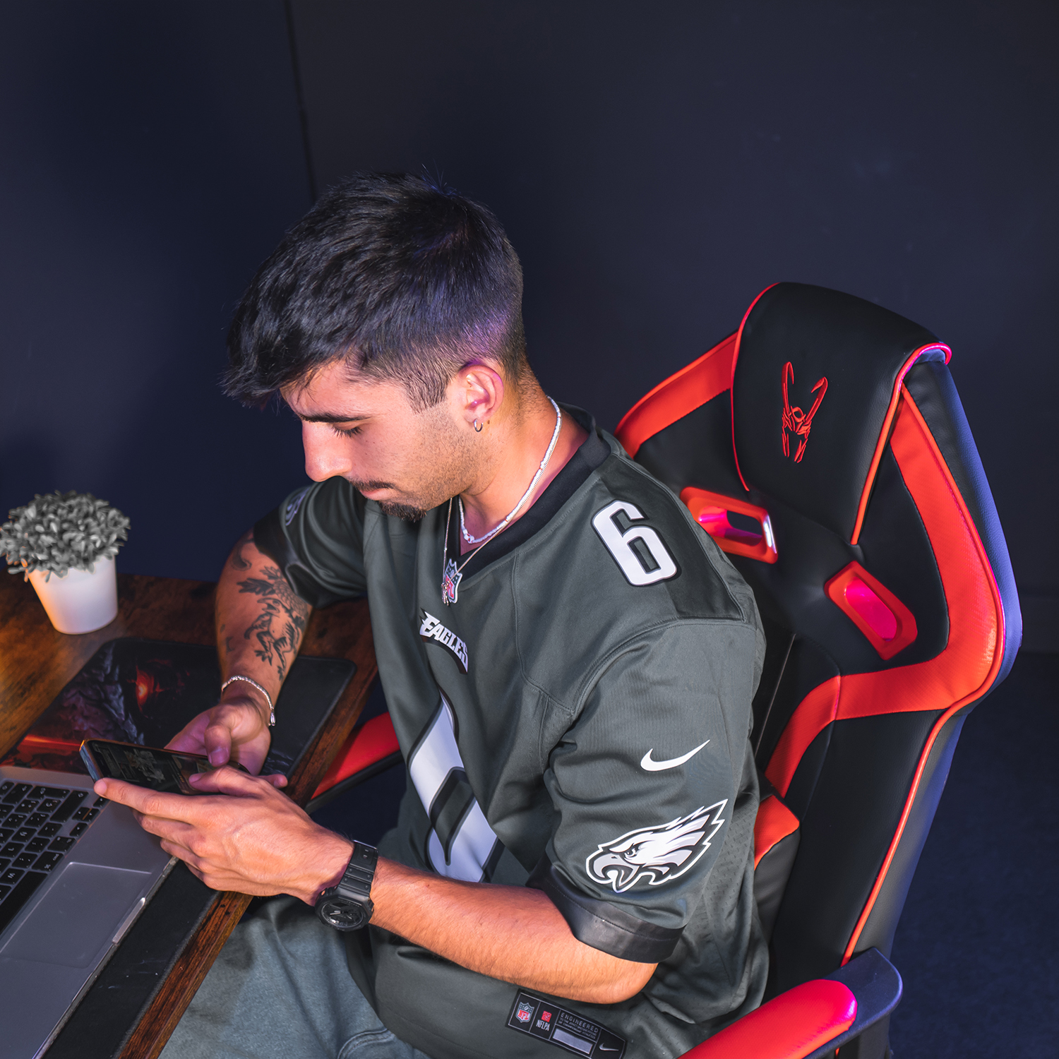 Un hombre está sentado en una silla de juego mirando su teléfono. Un portátil está en el escritorio.