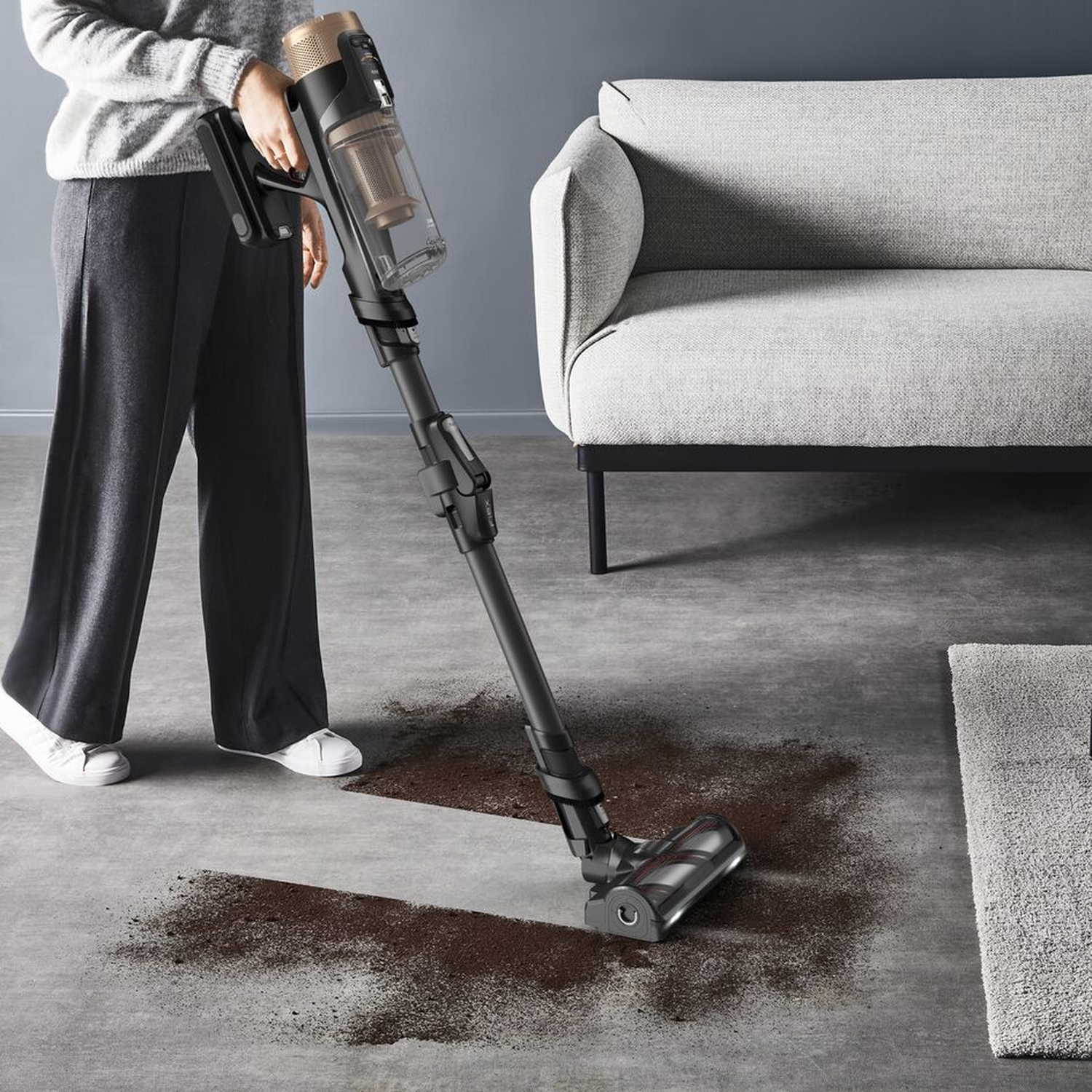 Une personne passe l'aspirateur sur un sol avec un aspirateur sans fil. De la saleté brune est visible.