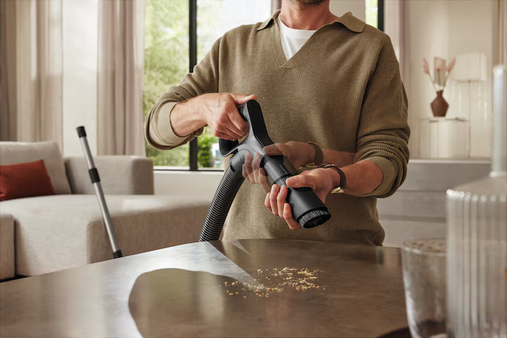 Mężczyzna czyści blat odkurzaczem. Brązowy blat. Beżowa sofa w tle.