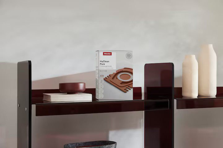 Półka z pudełkiem worków do odkurzacza, miską, książką i dwoma wazonami.