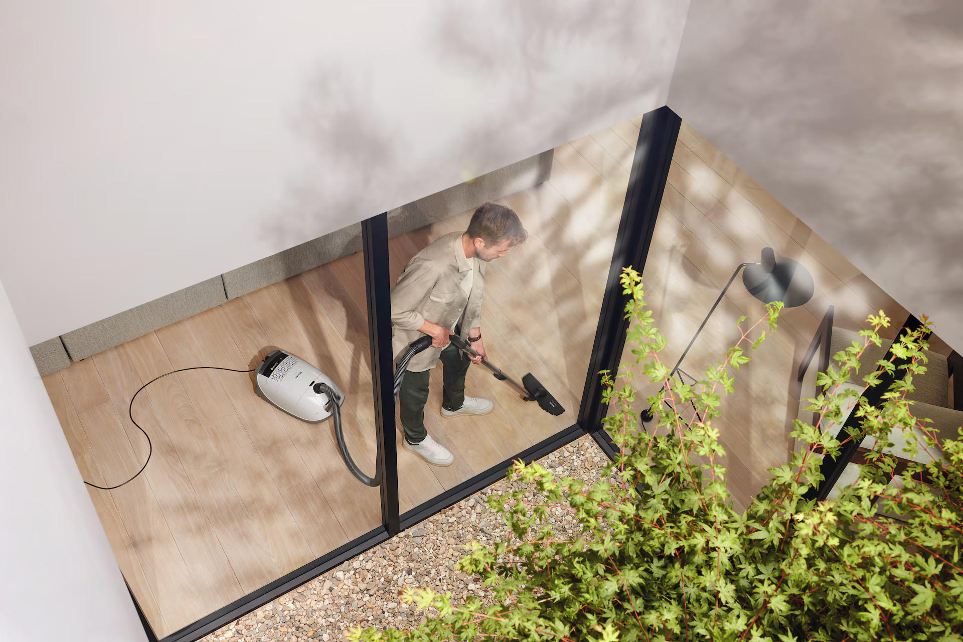 Mężczyzna odkurzający pokój z drewnianą podłogą. Odkurzacz jest biały. Rośliny otaczają pokój.