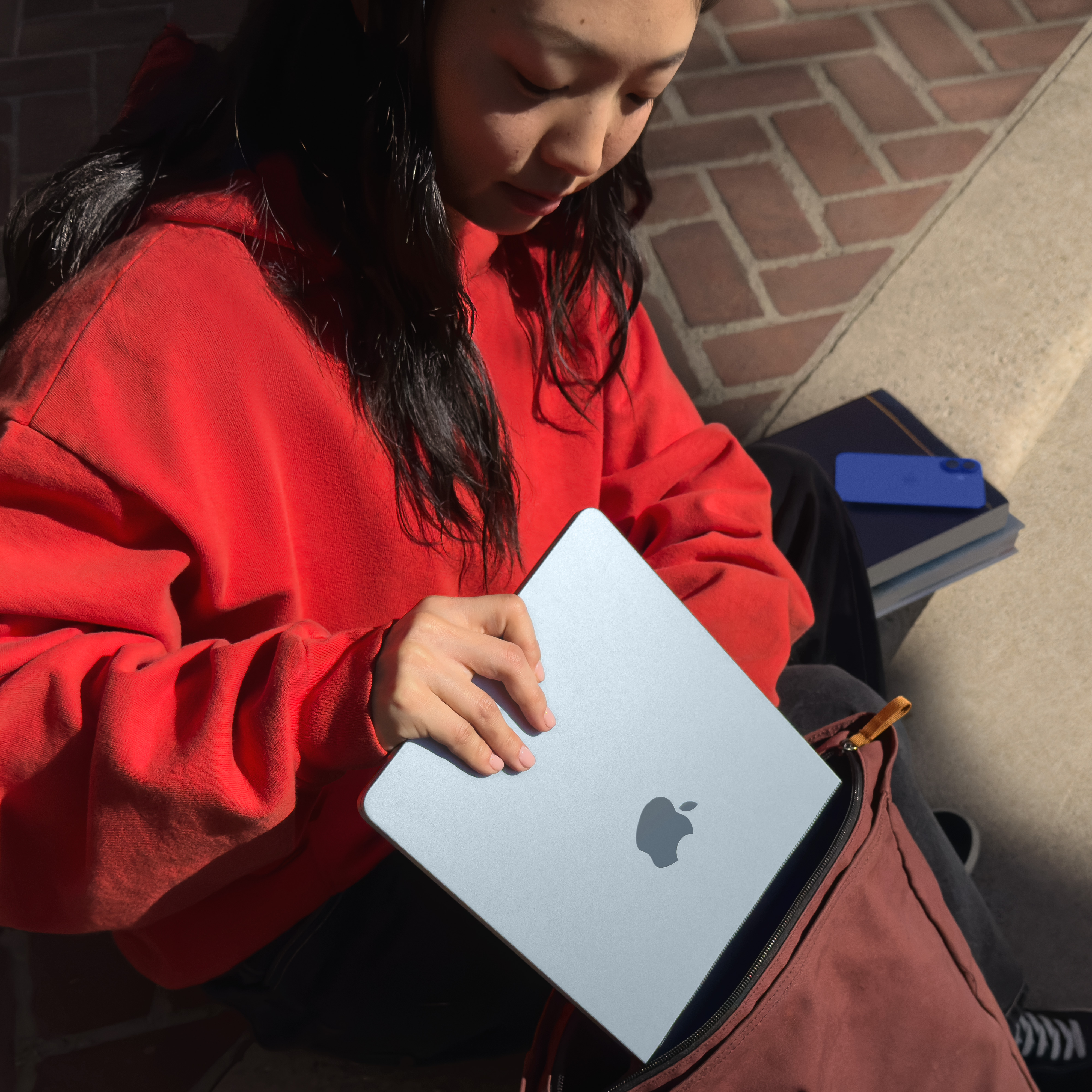 Eine Person in einem roten Kapuzenpullover hält einen blauen Laptop aus einem Rucksack. Ein Buch und ein Telefon sind in der Nähe.