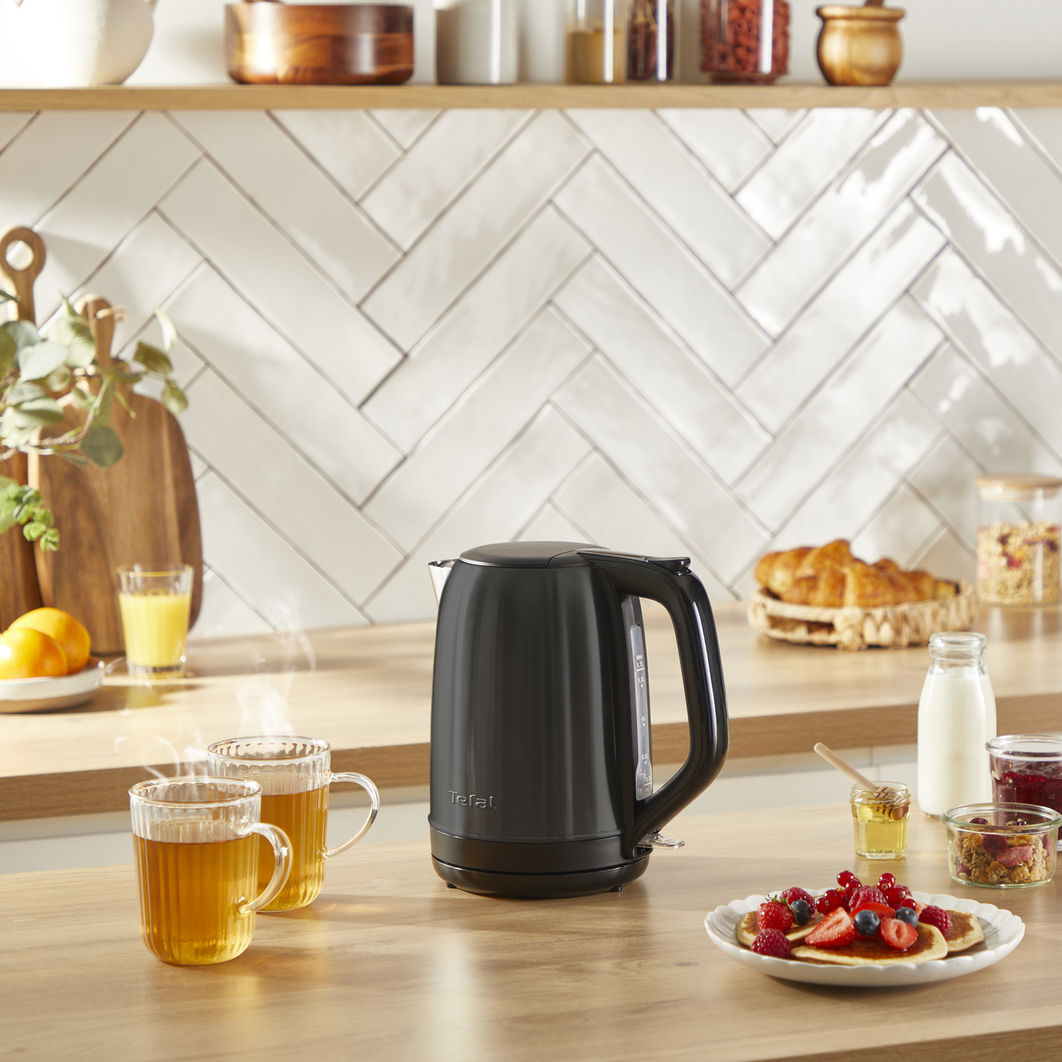 A black kettle on a wooden table with breakfast items and two glass cups of tea.