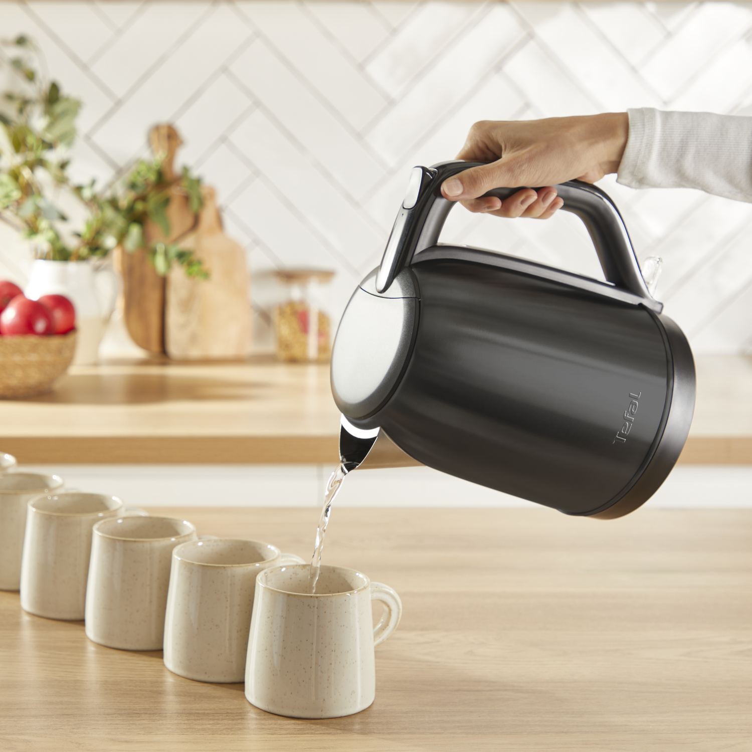 Person pouring water from a black kettle into mugs on a wooden surface.