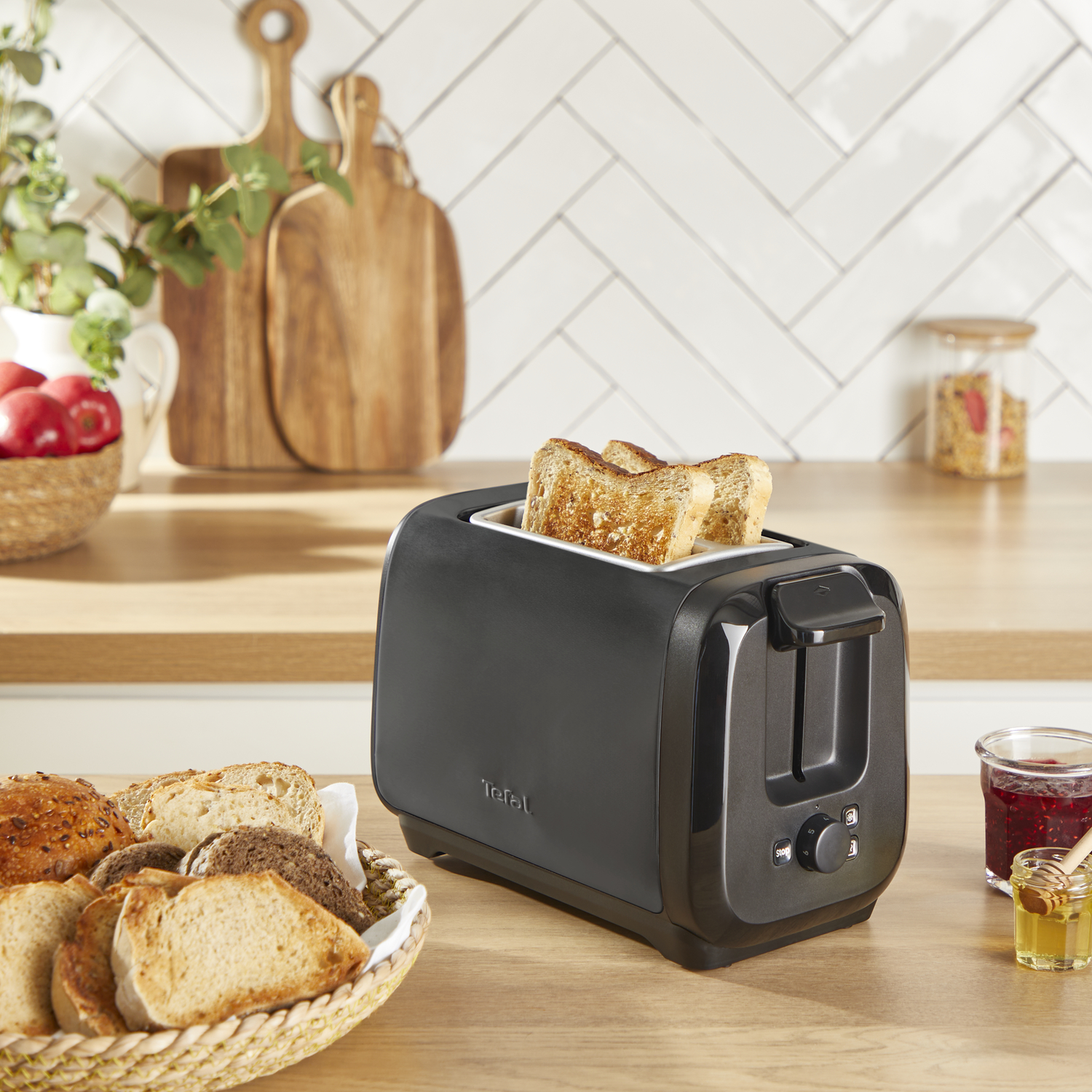 Zwarte broodrooster met twee sneetjes toast, omringd door brood en jam.