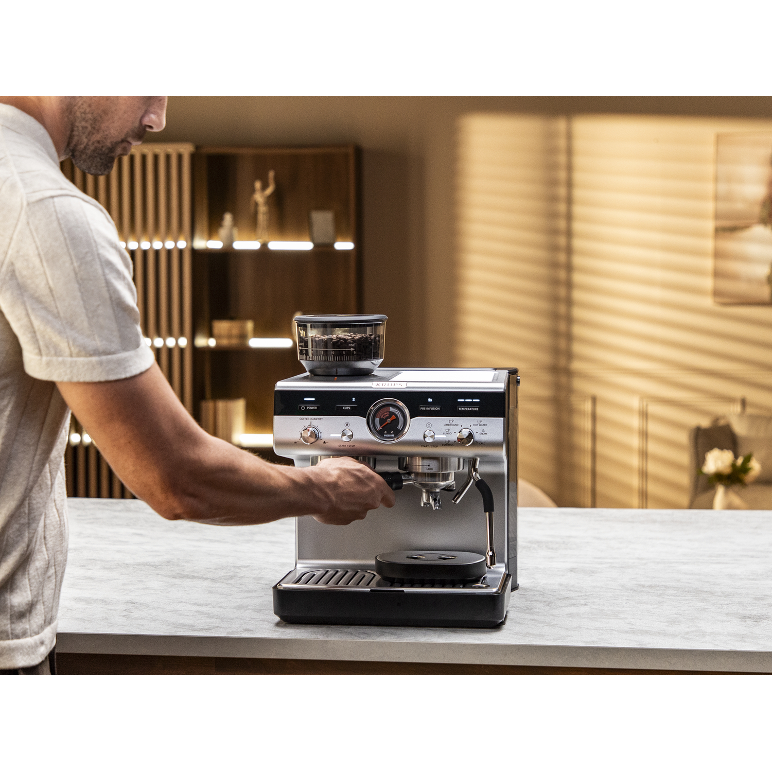 A man using a silver coffee machine with a grinder on top. The background is a neutral-toned kitchen.