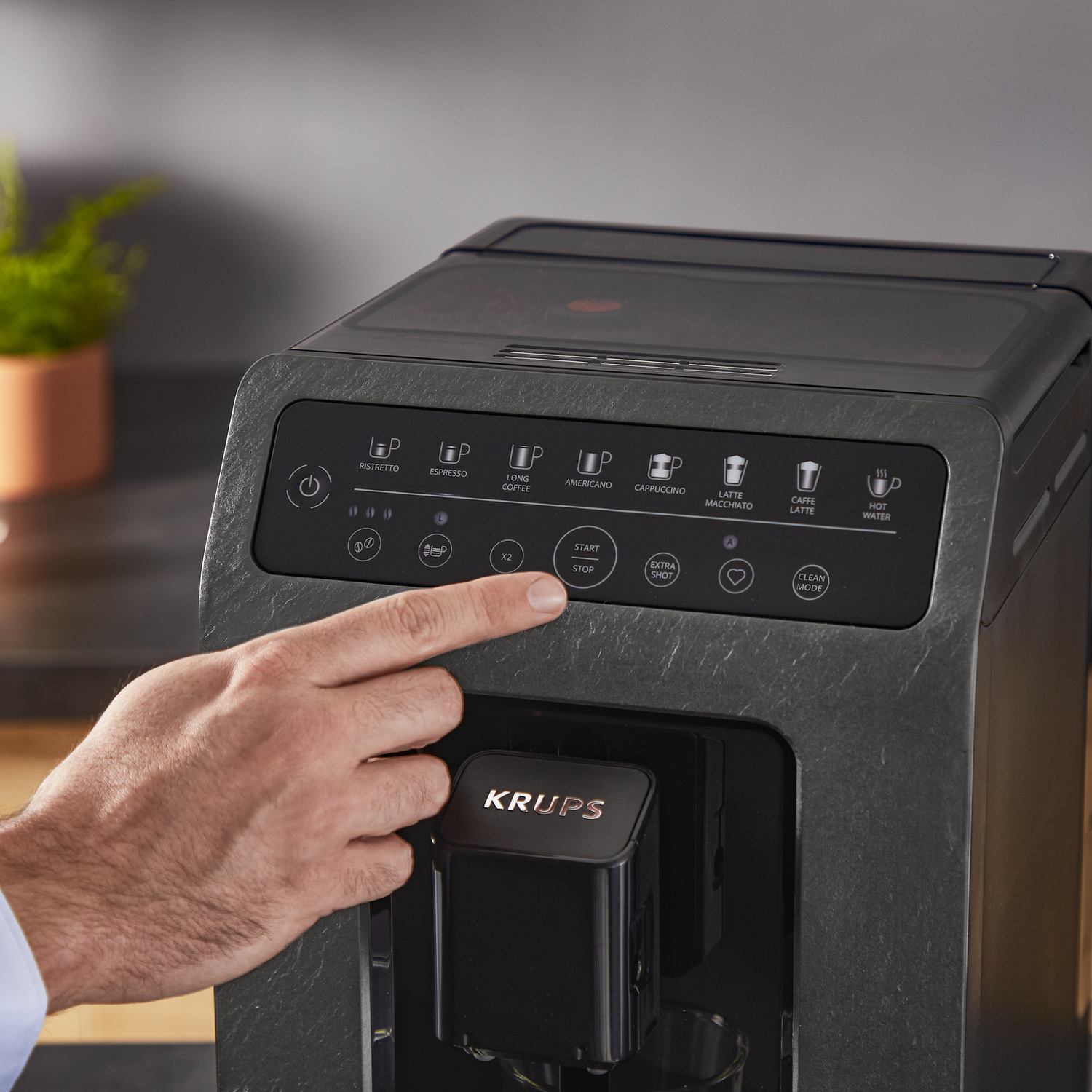 Een hand drukt op een knop op een donkergrijze koffiemachine. Het merk is KRUPS.
