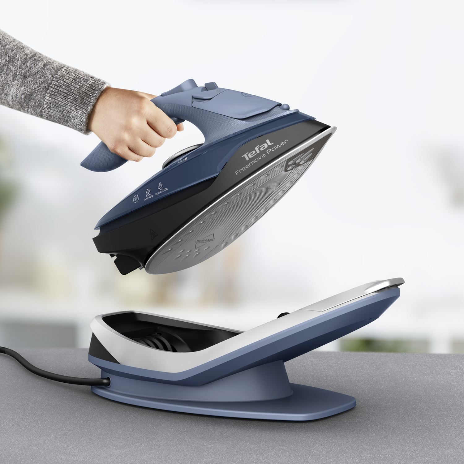 A hand holding a blue iron above its charging station on a gray surface. The background is blurred.