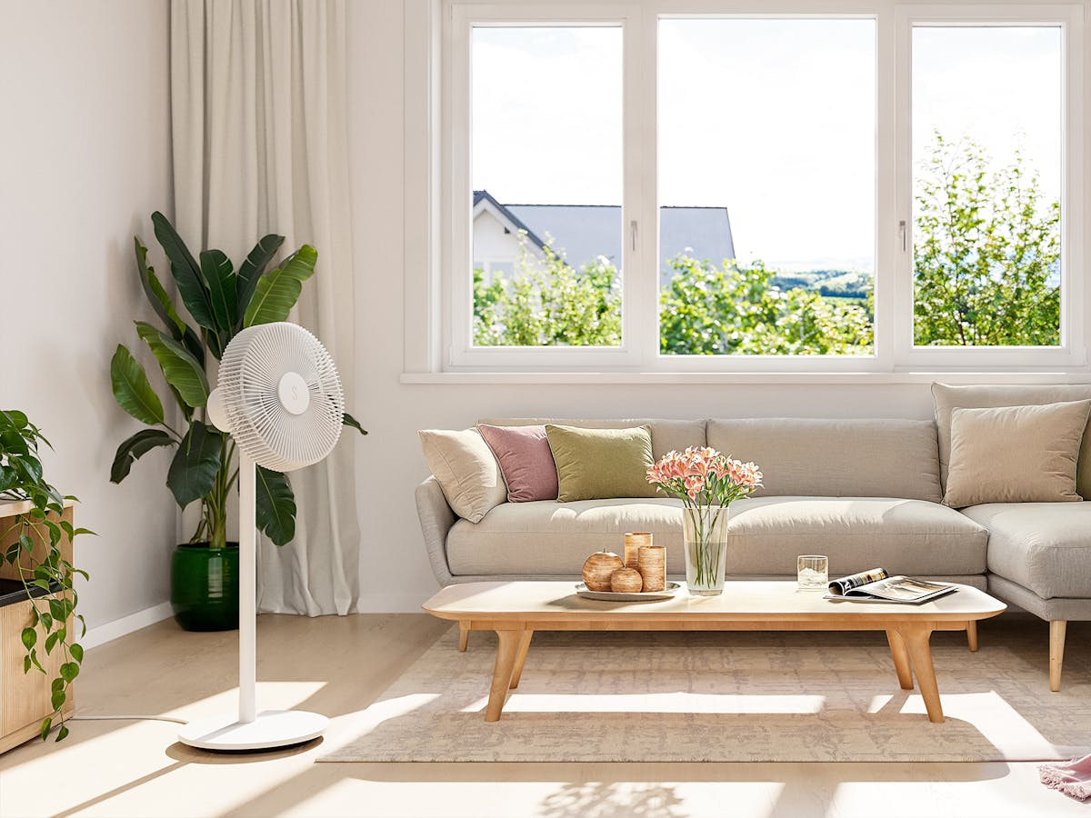 Ein Wohnzimmer mit einem weißen Ventilator, Sofa, Tisch, Pflanzen und einem Fenster.