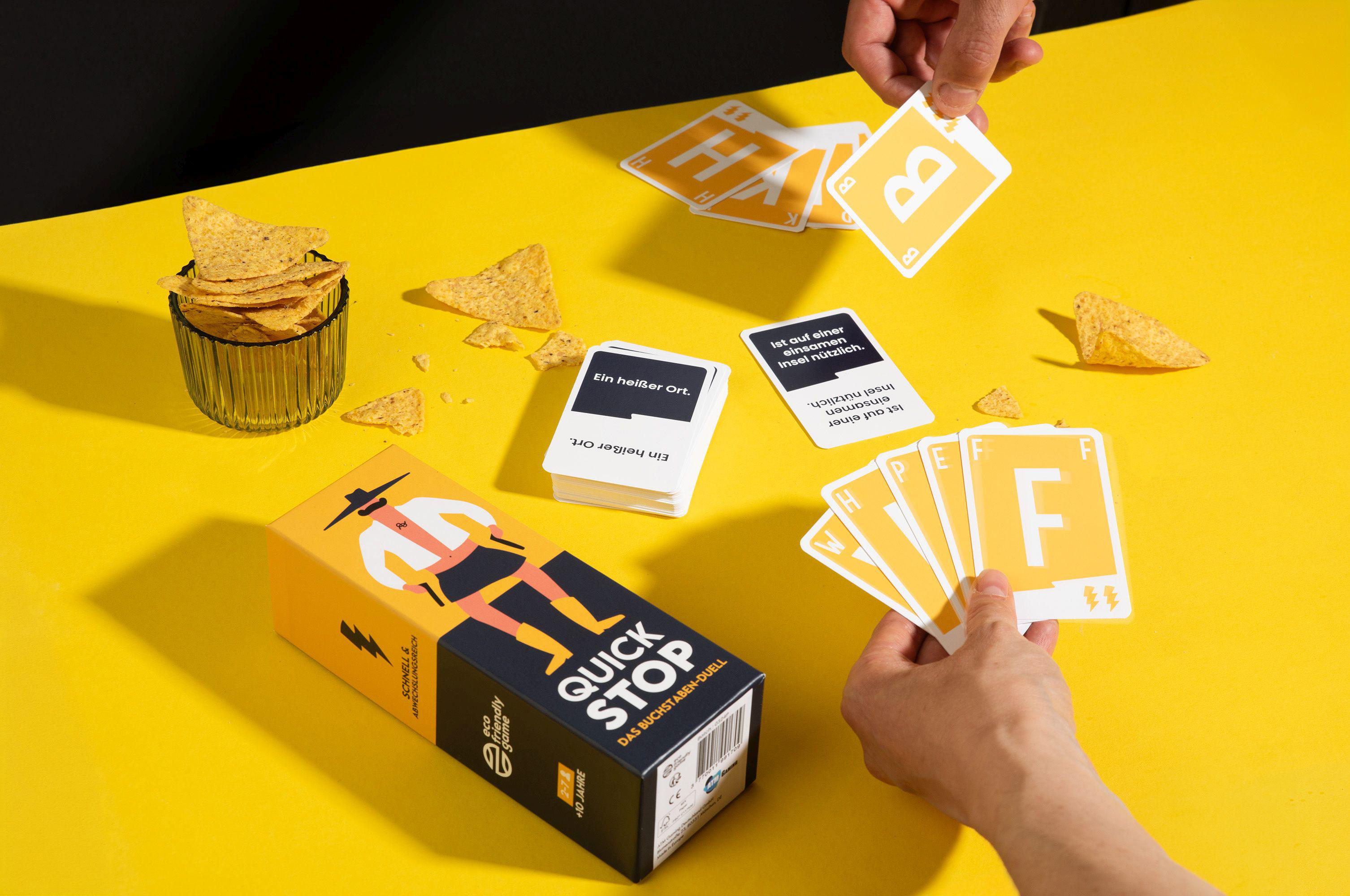 Ein Brettspiel-Setup mit Spielkarten, Chips und einer Box auf gelber Oberfläche. Zwei Hände sind sichtbar.