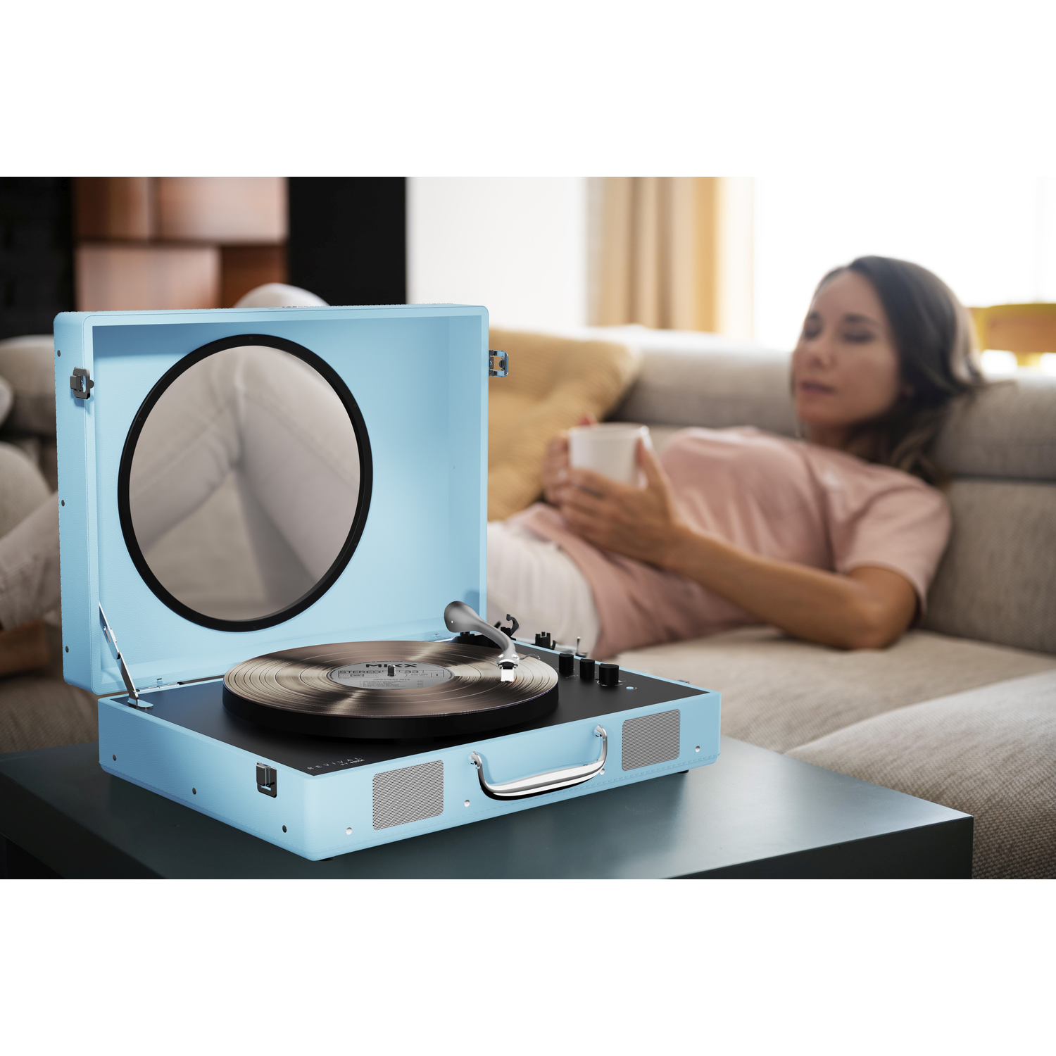A light blue record player case with a vinyl record and needle. A woman lies on a sofa in the background.