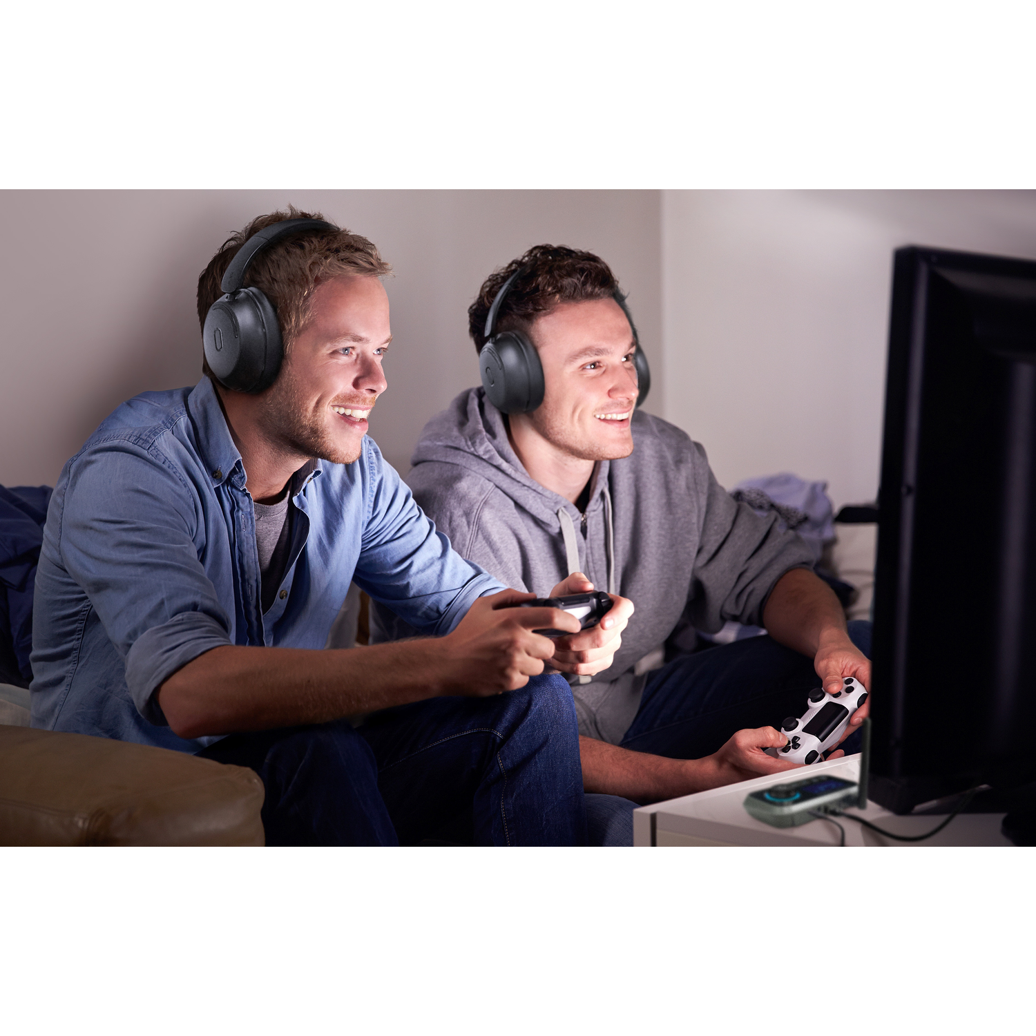 Two young men wearing headphones play video games, smiling, holding controllers, and watching a TV. One is in denim, the other in a grey hoodie.