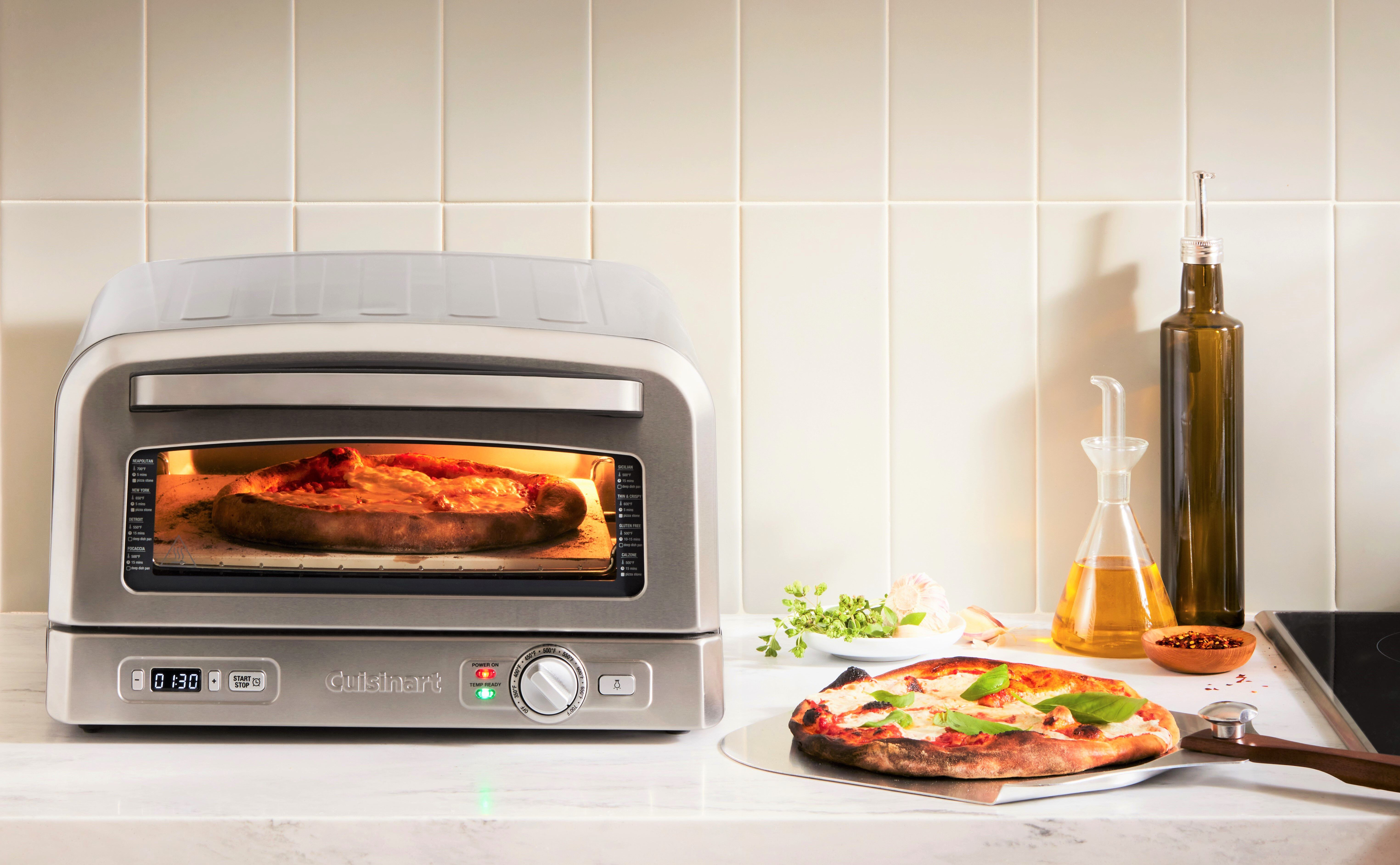 Une pizza cuit dans un four argenté, avec une autre pizza sur le comptoir.