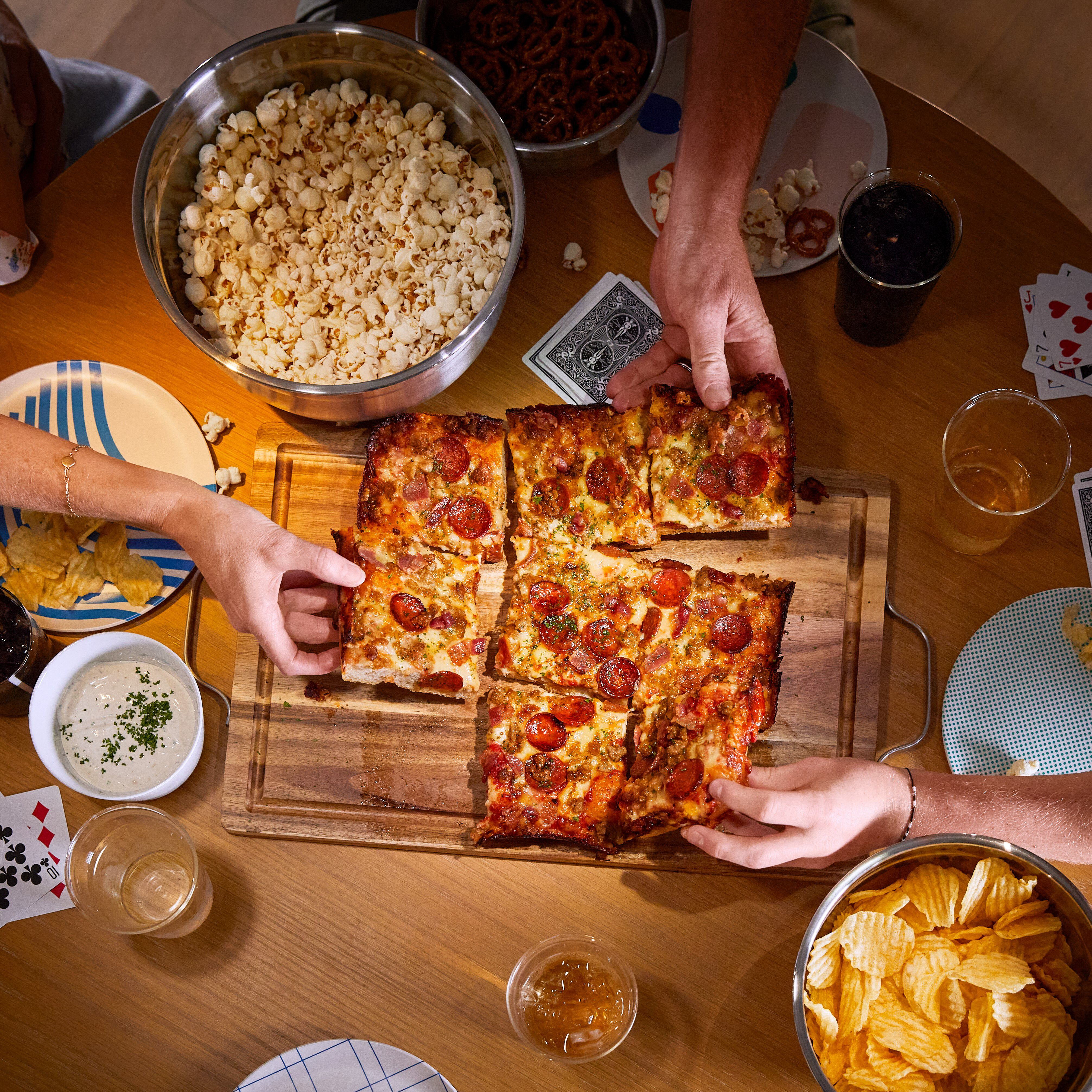 Vue aérienne de personnes partageant une table avec de la pizza, du pop-corn, des chips et des boissons. Mains atteignant la pizza.