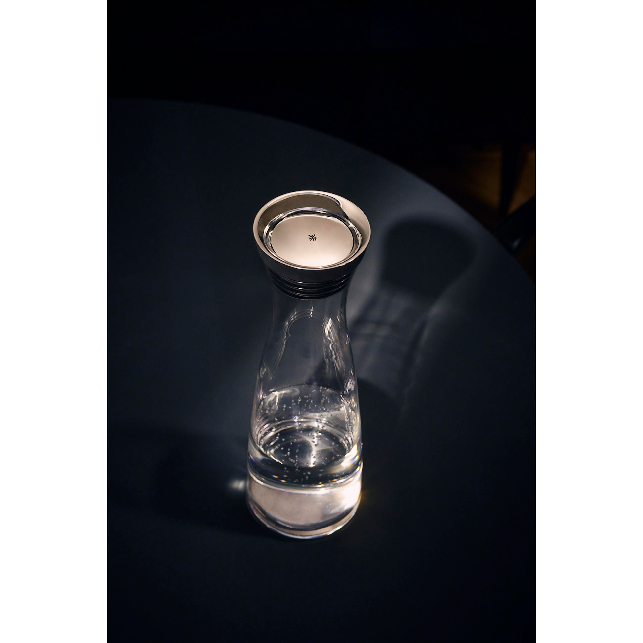Eine Glaskaraffe mit Wasser, mit silbernem Deckel, auf dunkler Oberfläche.