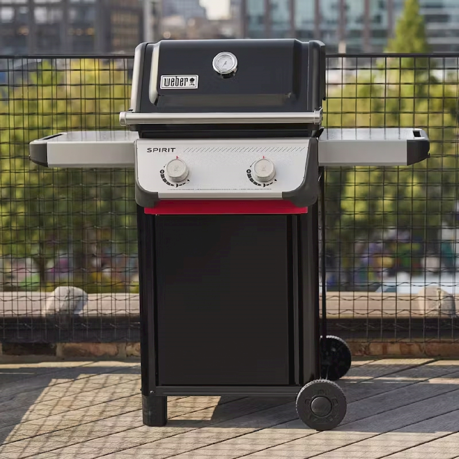 Un barbecue Weber nero con un accento rosso, su un ponte di legno. Una recinzione metallica sullo sfondo.