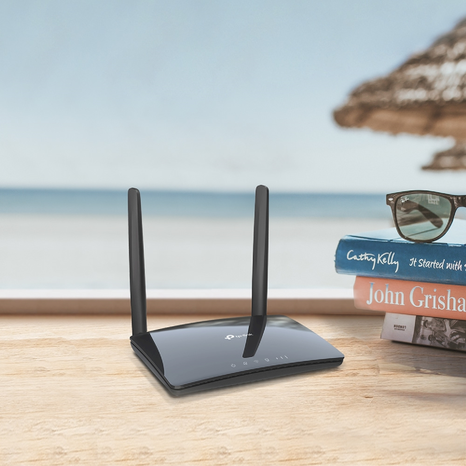 Un router nero con antenne è appoggiato su un tavolo con libri e occhiali da sole, sfondo spiaggia.