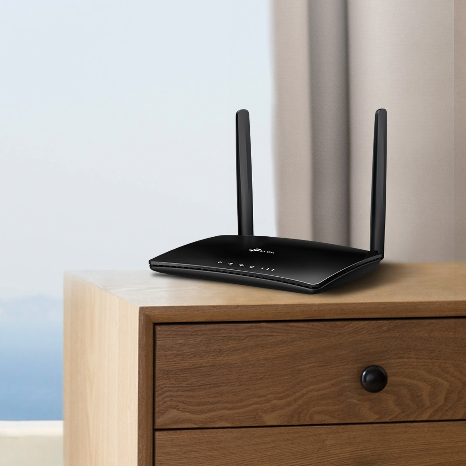 Un router nero con due antenne è posizionato su un comodino di legno vicino a una finestra.