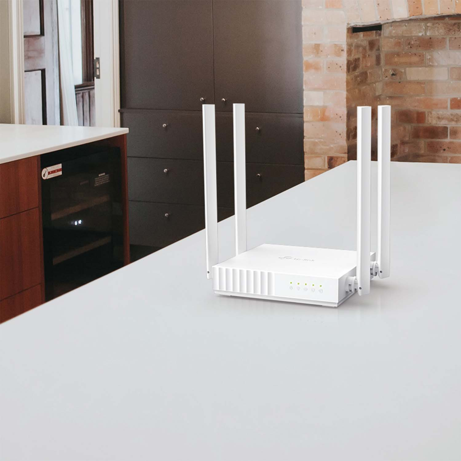 Un router bianco con quattro antenne posizionato su un piano di lavoro bianco vicino a un mobile scuro e un muro di mattoni.
