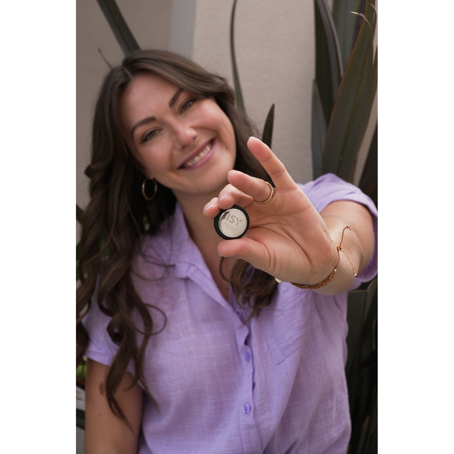 A woman with long brown hair smiles, holding a small makeup product in her hand.