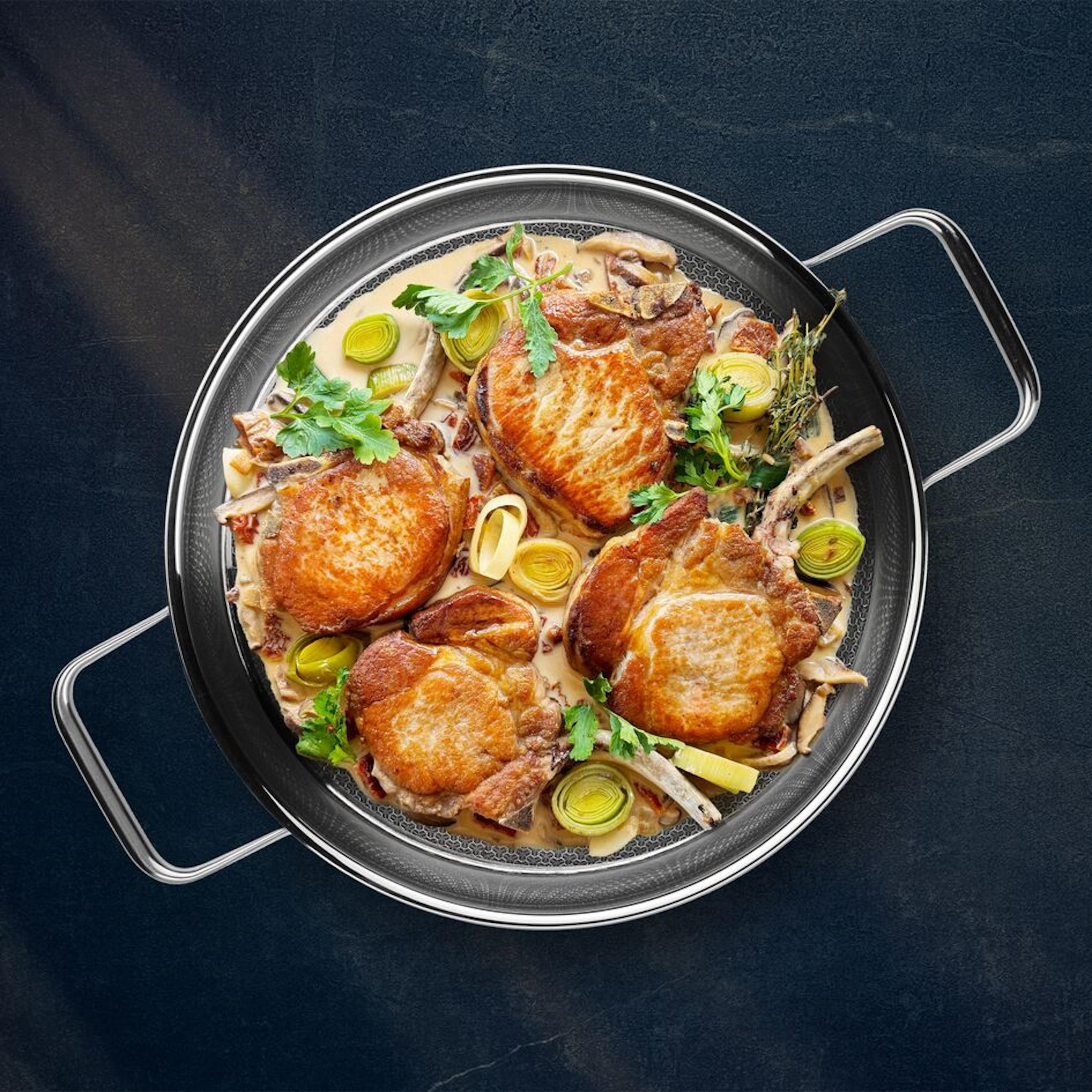 Draufsicht auf gekochte Schweinekoteletts mit Gemüse und Soße, in einer Pfanne auf dunkler Oberfläche.