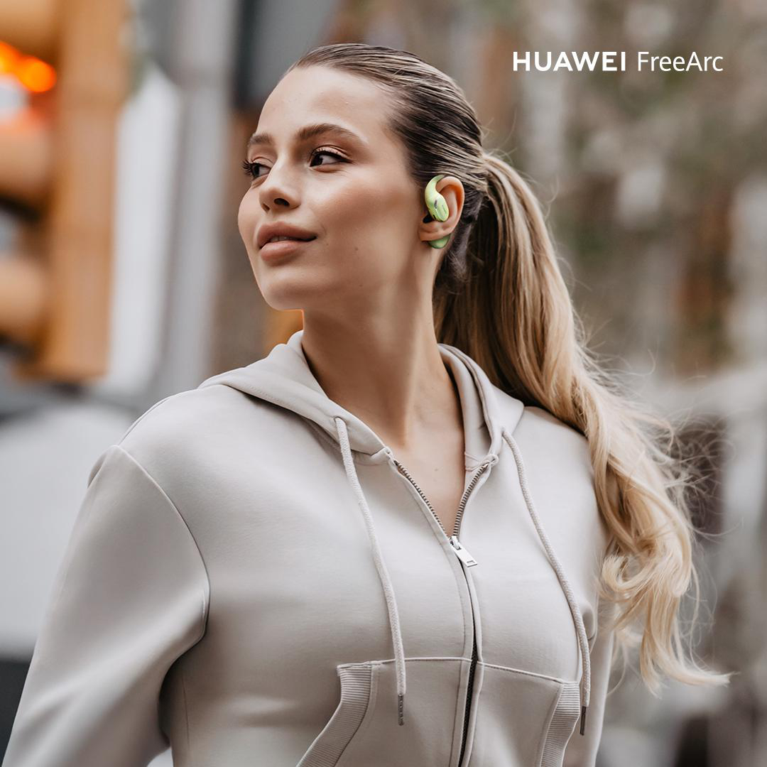 Eine Frau mit blonden Haaren und einem Kapuzenpullover, die Ohrhörer trägt. Der Hintergrund ist verschwommen, mit einer Ampel sichtbar.