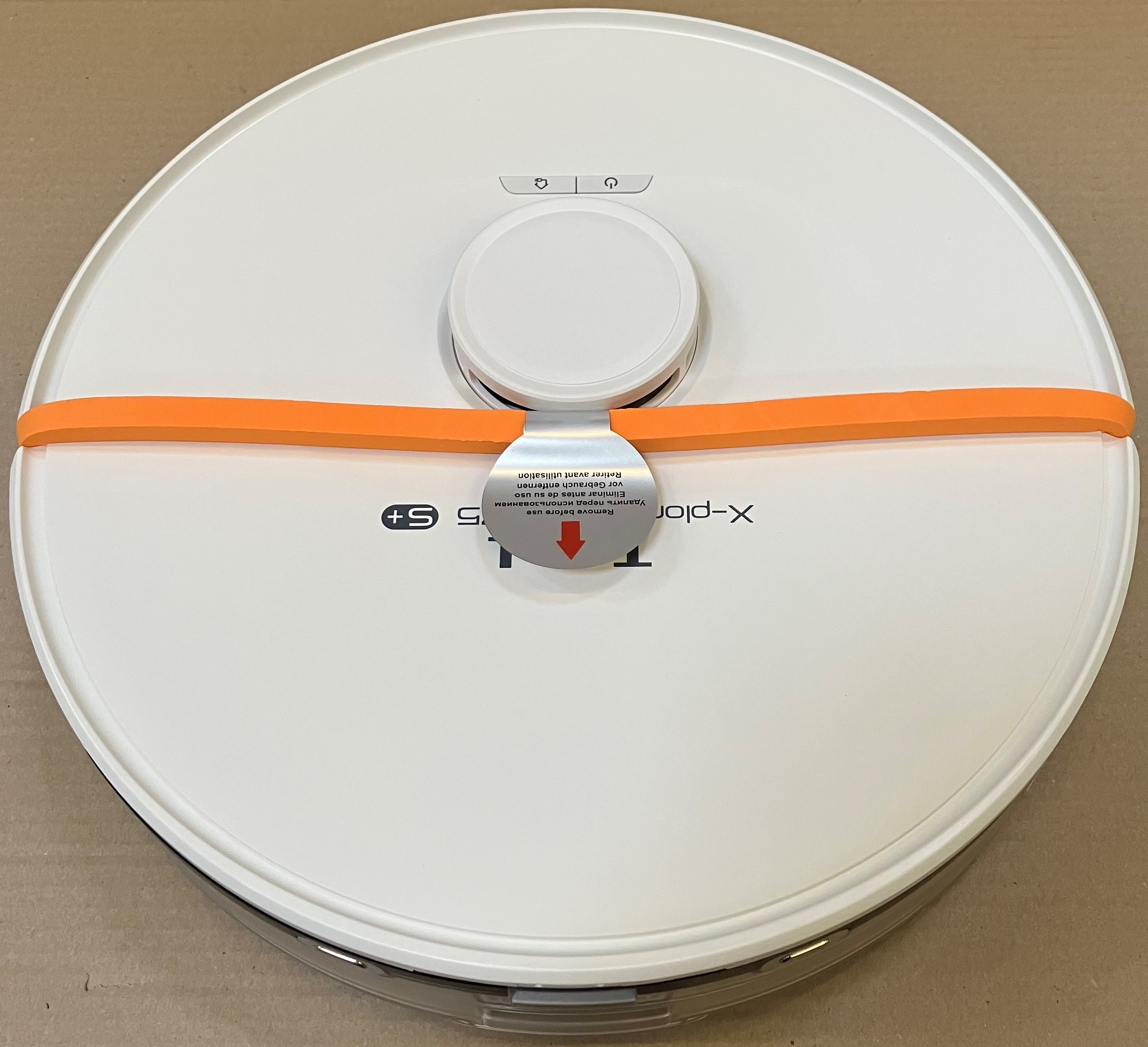 Biały odkurzacz automatyczny z pomarańczową opaską. Widoczne przyciski i tekst. Umieszczony na jasnobrązowej powierzchni.