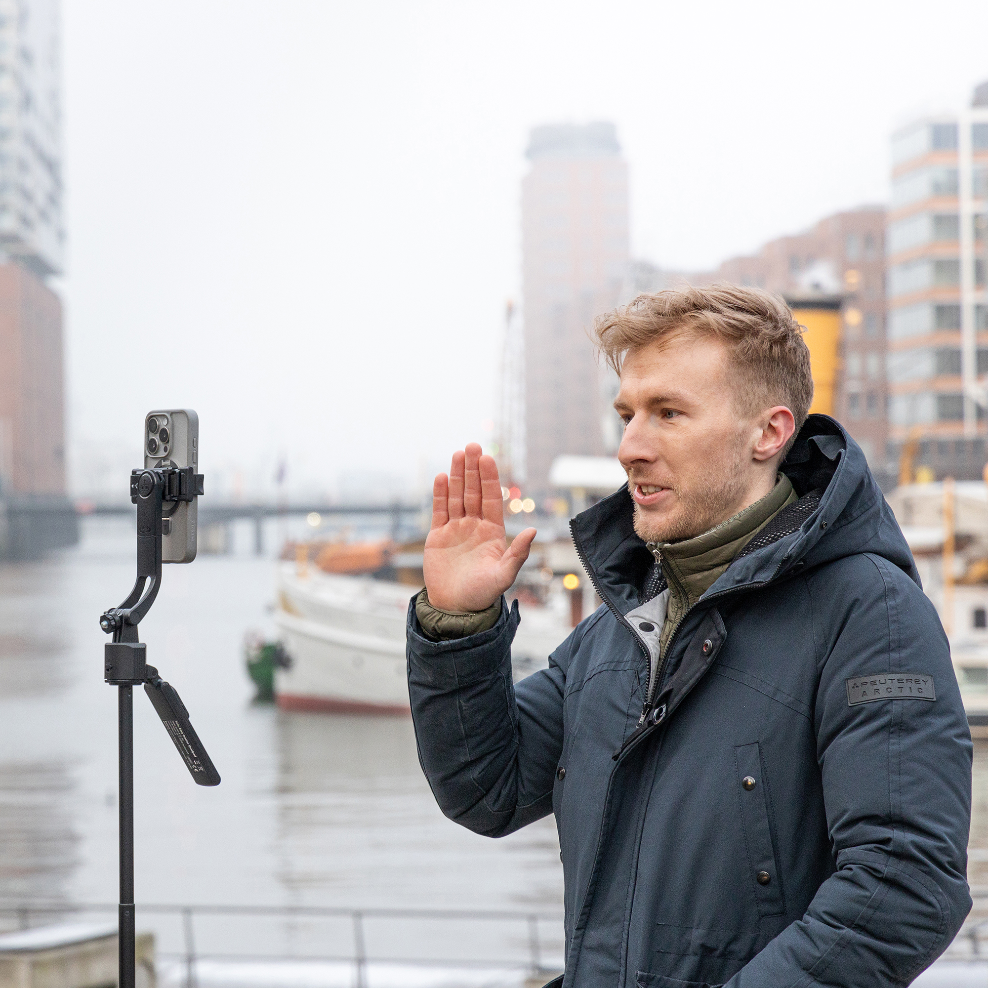 Ein Mann steht am Wasser, hält ein Telefon auf einem Stativ und macht eine Geste mit der Hand.