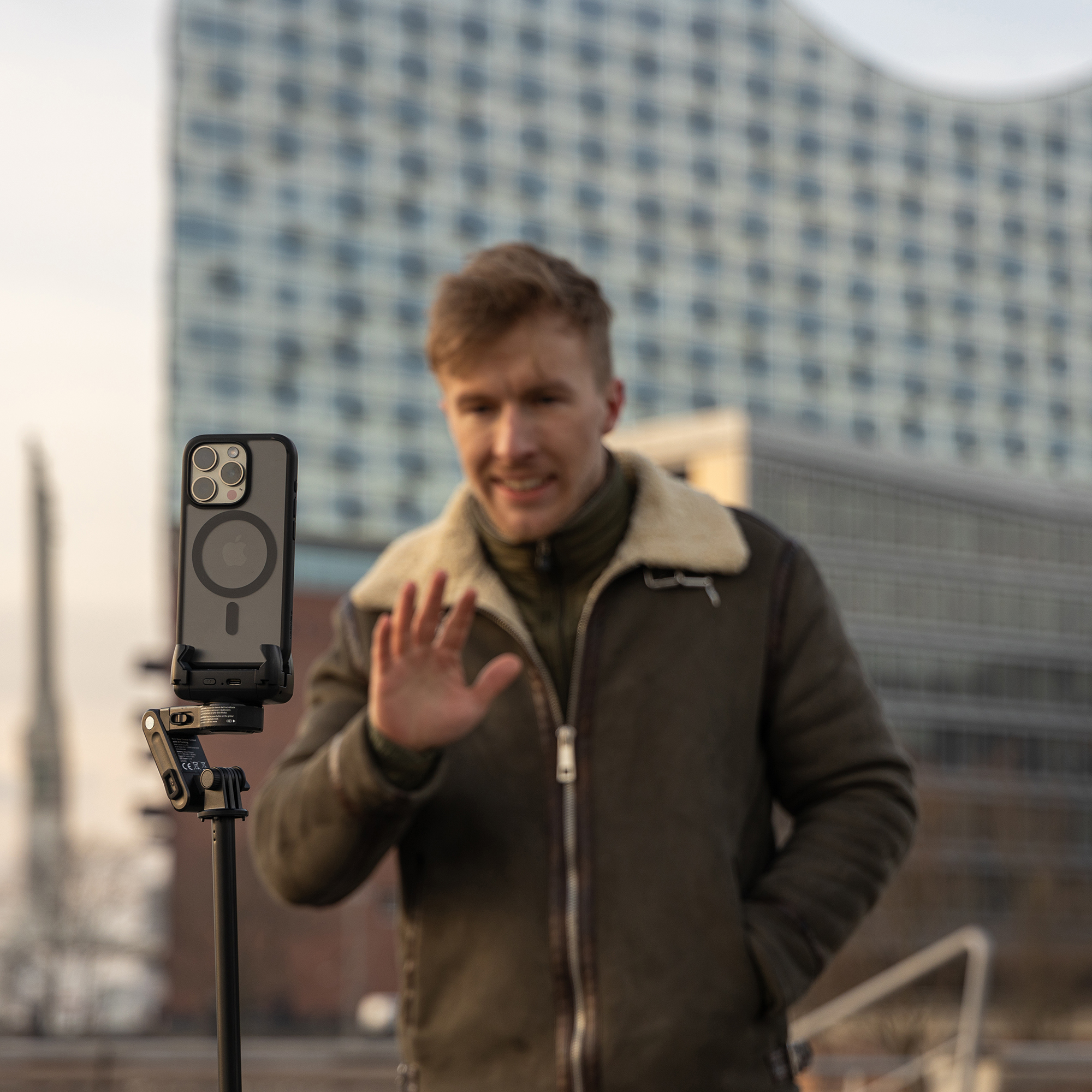 Homme tenant un téléphone sur une perche à selfie. Bâtiments en arrière-plan. L'homme salue.