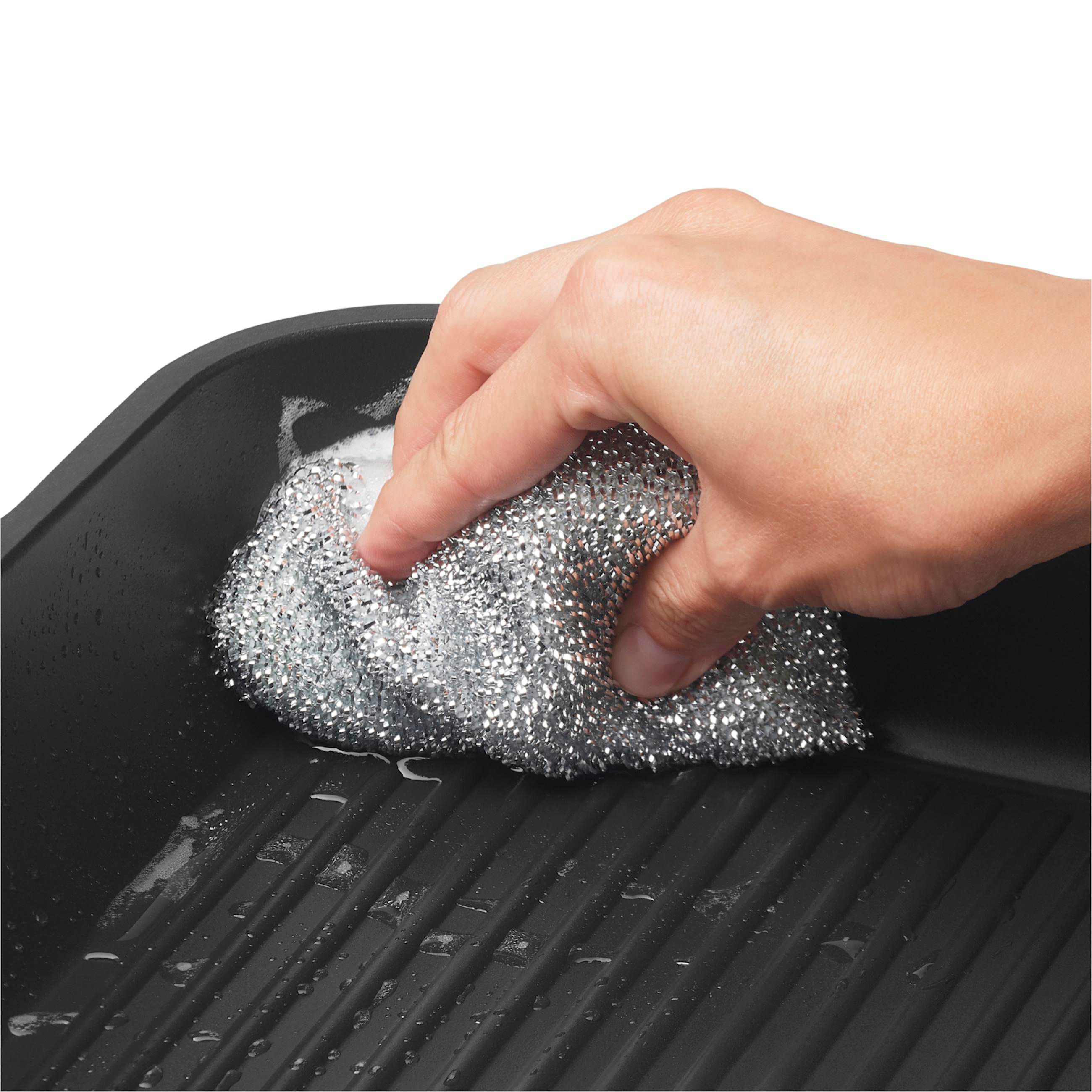 Hand reinigt eine schwarze Grillpfanne mit einem silbernen Schwamm. Nahaufnahme.