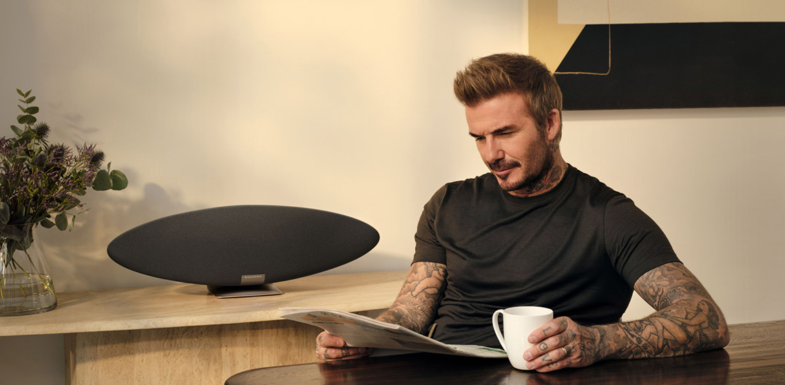 David Beckham czyta gazetę, trzyma filiżankę, głośnik na stole.