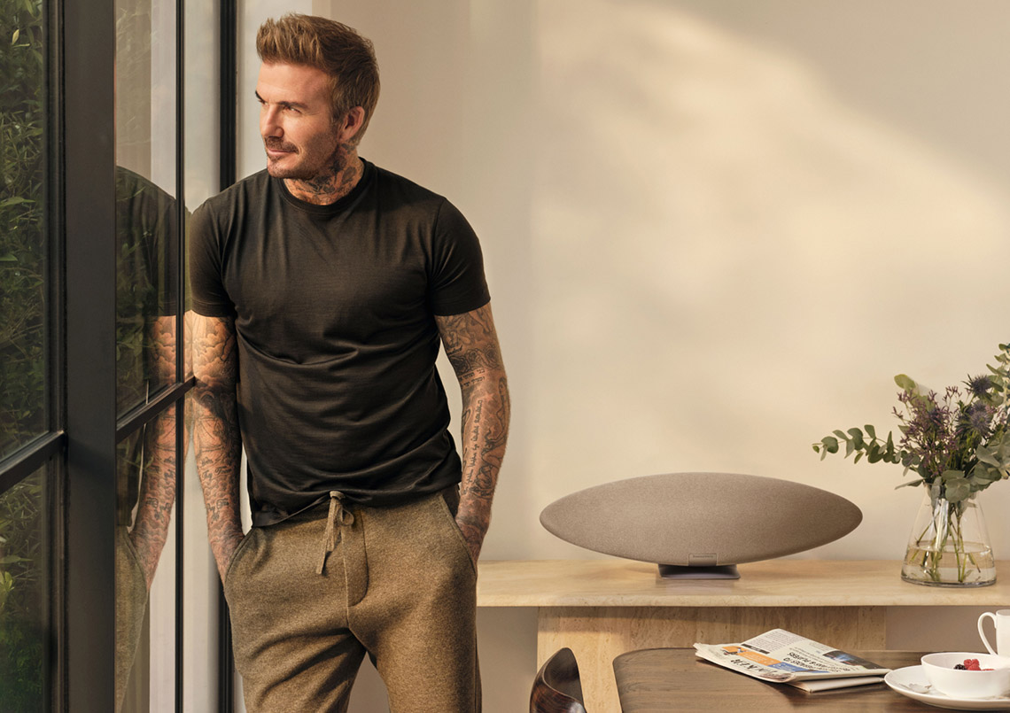 David Beckham w pokoju, patrzący przez okno. Na stole głośnik i kwiaty.