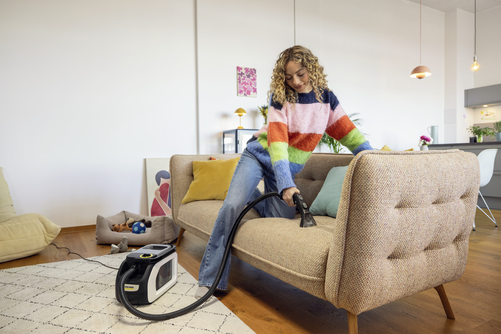 Kobieta czyszcząca kanapę odkurzaczem do dywanów. W tle legowisko dla psa i zabawki.