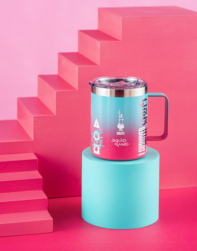 Eine blau-rosa Tasse mit Text steht auf einem blauen Podest. Im Hintergrund eine pinke Treppe.